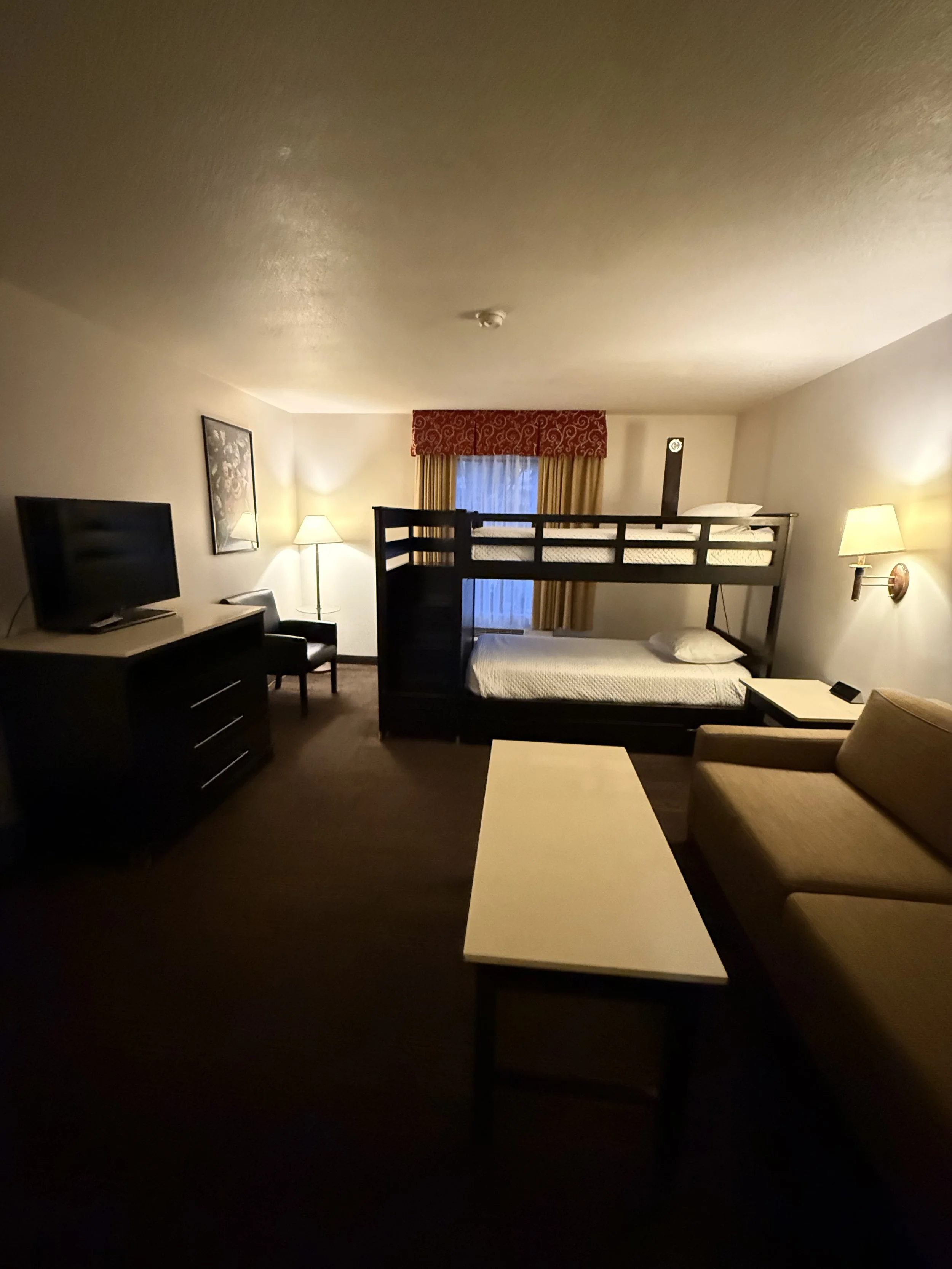 Hotel room with a bunk bed, a TV, a lamp, a bed, a chair, a small table, and a window with curtains.