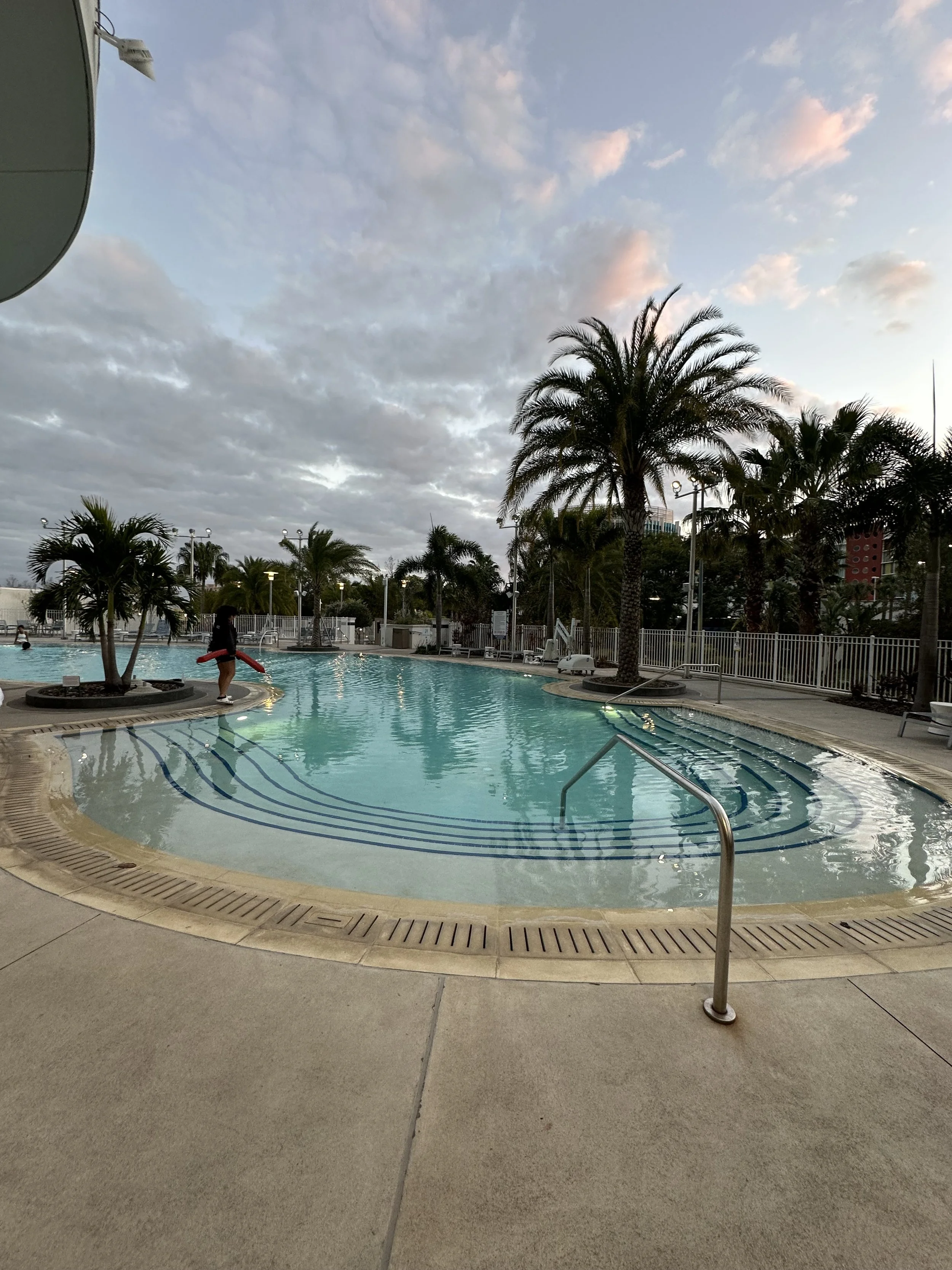 Empty outdoor swimming pool with steps and handrail, surrounded by palm trees and deck chairs, under a cloudy sky in the early evening.