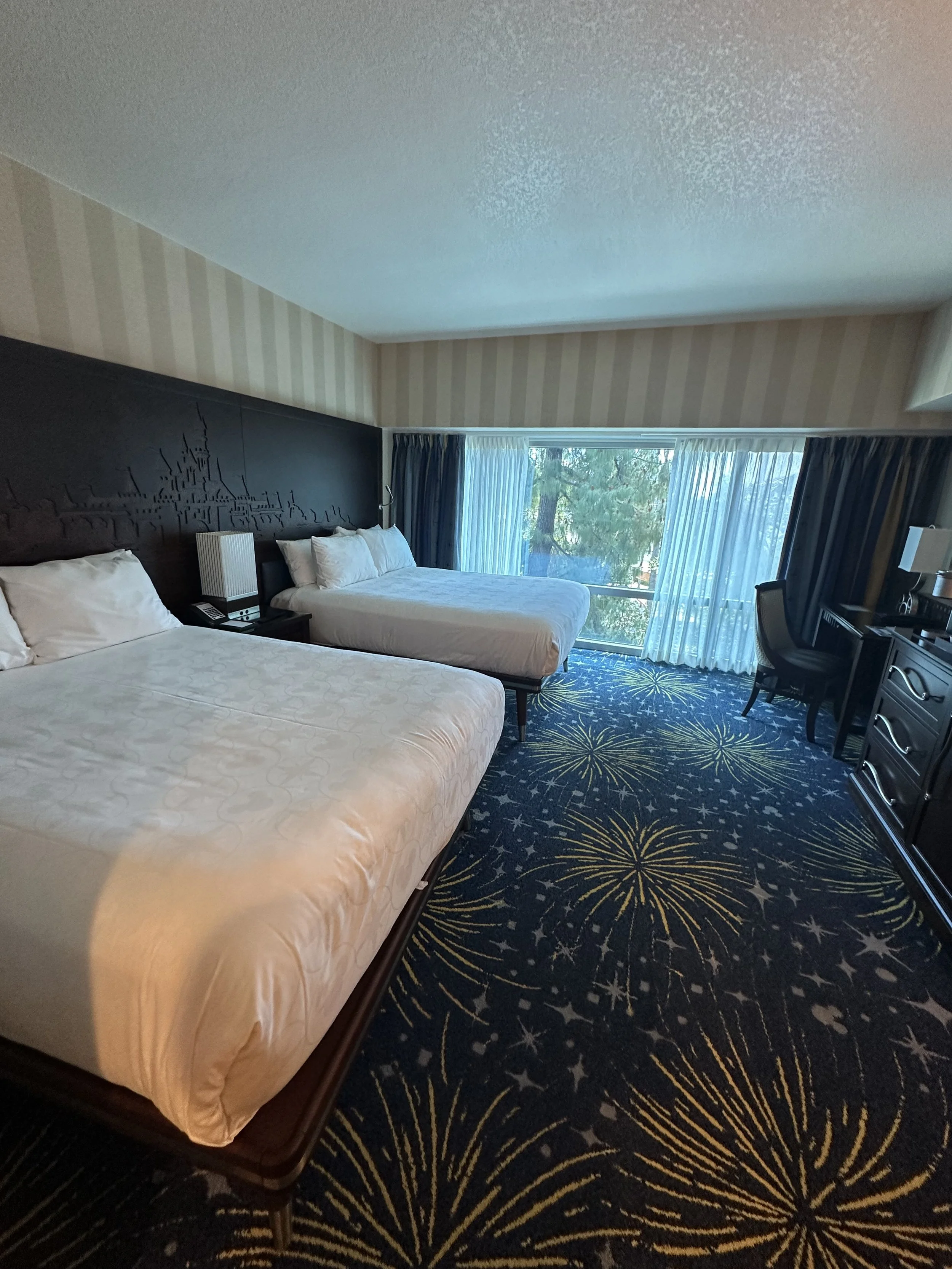 Hotel room with two beds, a large window with blue curtains, a desk, and a chair.