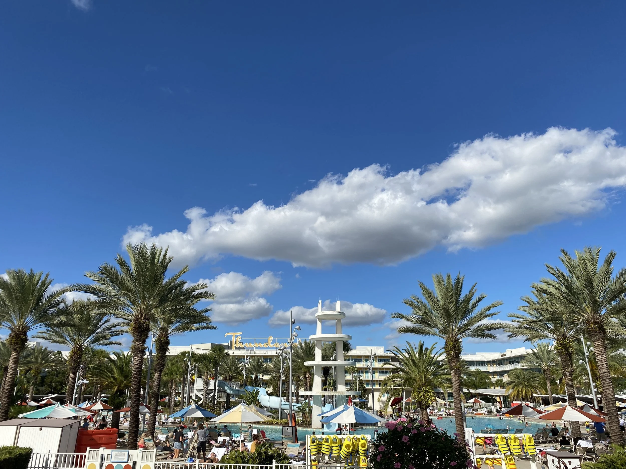 A busy tropical resort pool area with numerous sun loungers under umbrellas, tall palm trees, a white water slide structure, and a multi-story hotel building in the background under a bright blue sky with white clouds.
