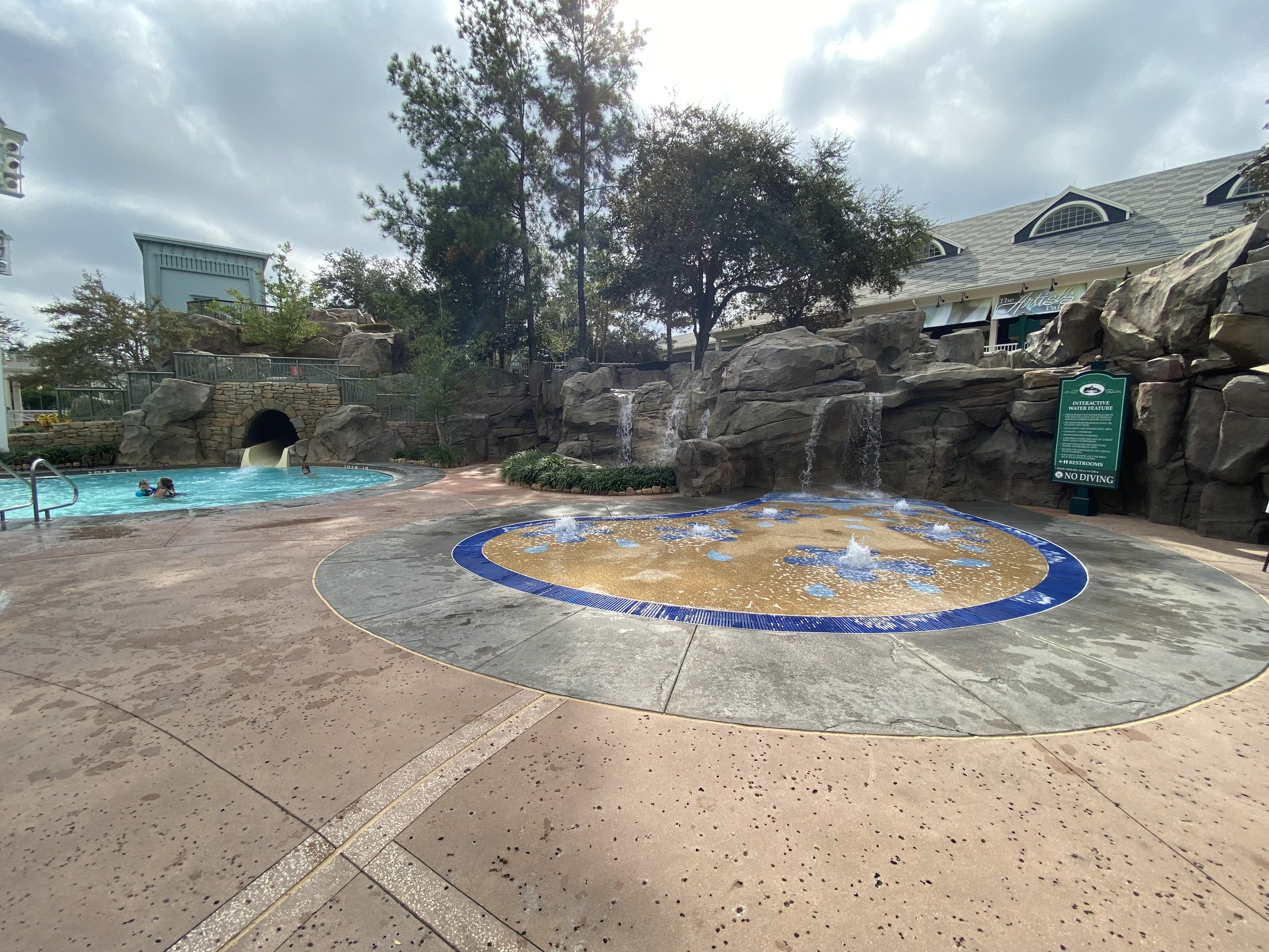 An outdoor pool area with a small waterslide, rocks, and water features. Two children are playing in the pool. The sky is cloudy, and there are trees and a building in the background.