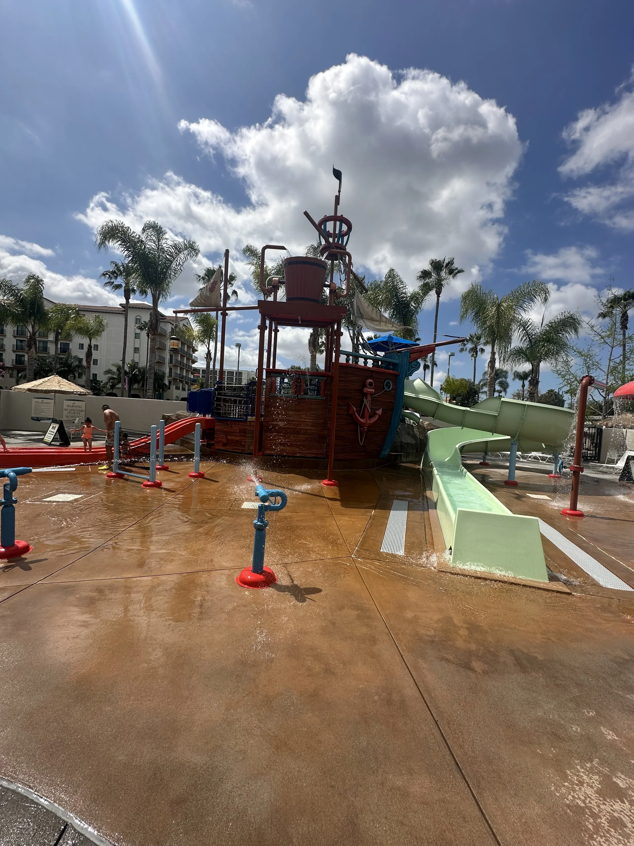 Water play area with a ship-shaped water slide, small fountains, and a splash pad under a partly cloudy sky with palm trees and buildings in the background.
