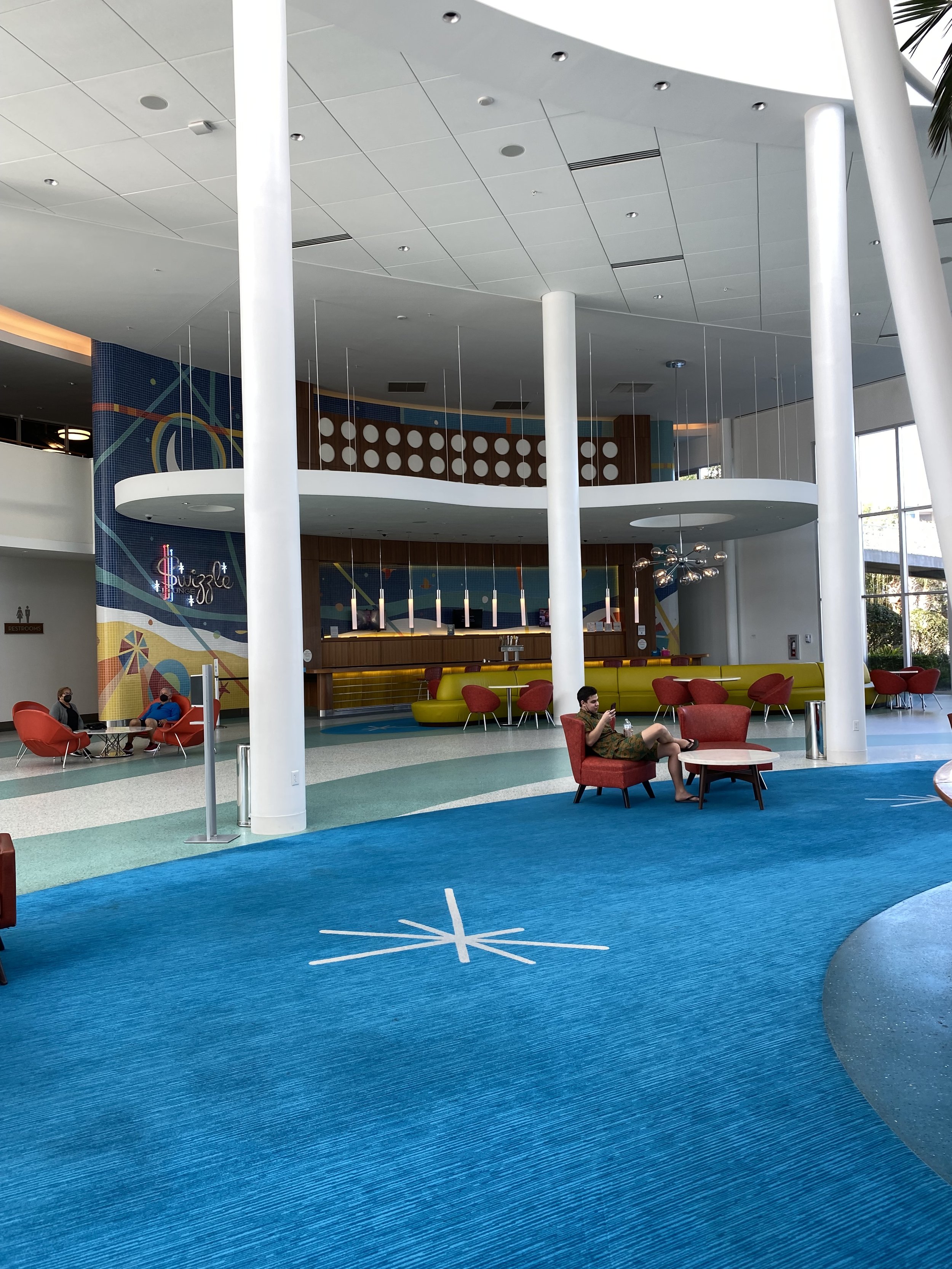 Modern indoor lounge area with colorful furniture, blue carpet, and large white columns. There are a few people seated, some using their phones, and a brightly decorated wall with a neon sign that says 'Shuffle.'