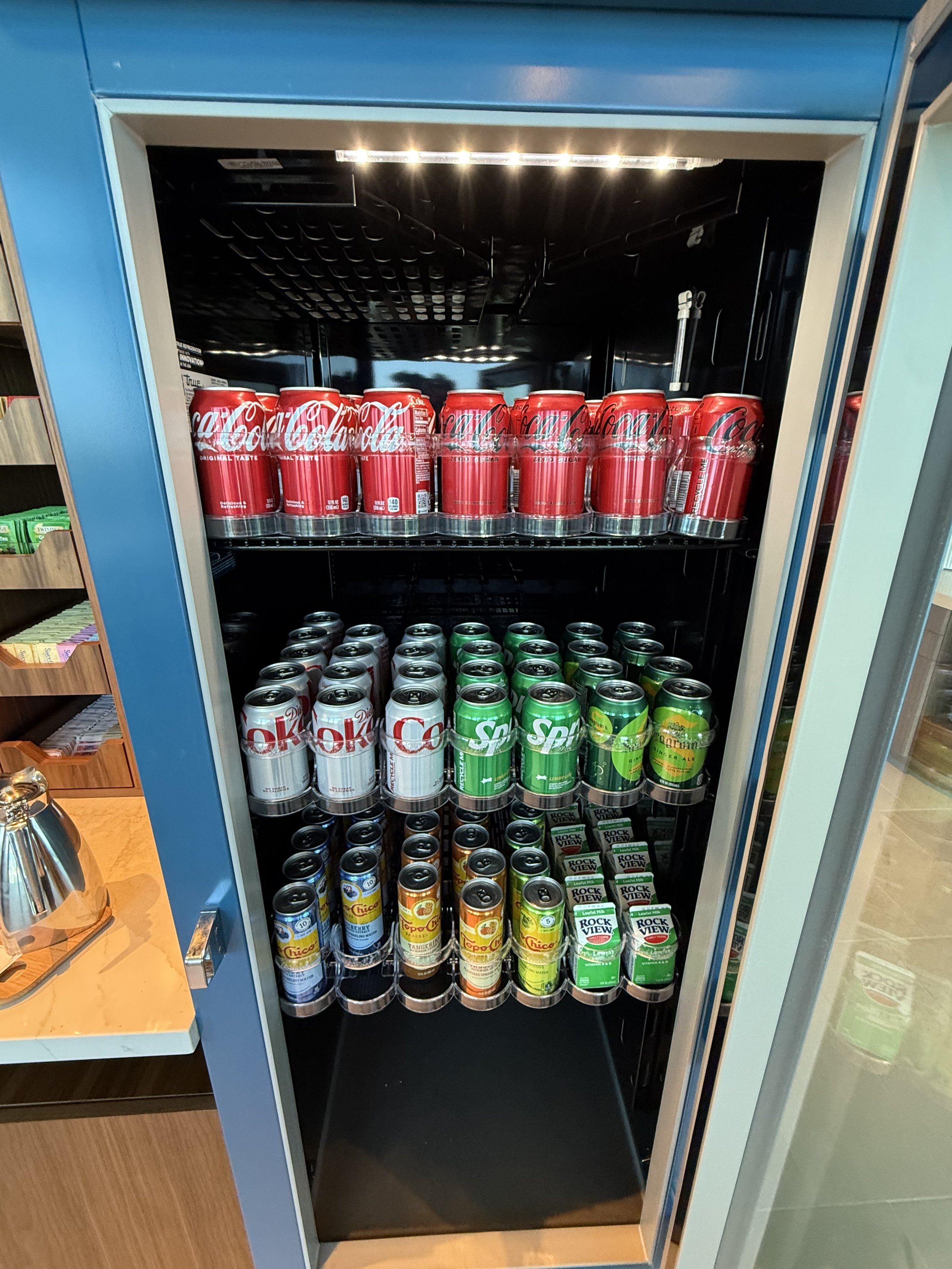 A vending machine with Coca-Cola cans on the top shelf, Sprite cans on the middle shelf, and various other canned beverages on the bottom shelf.