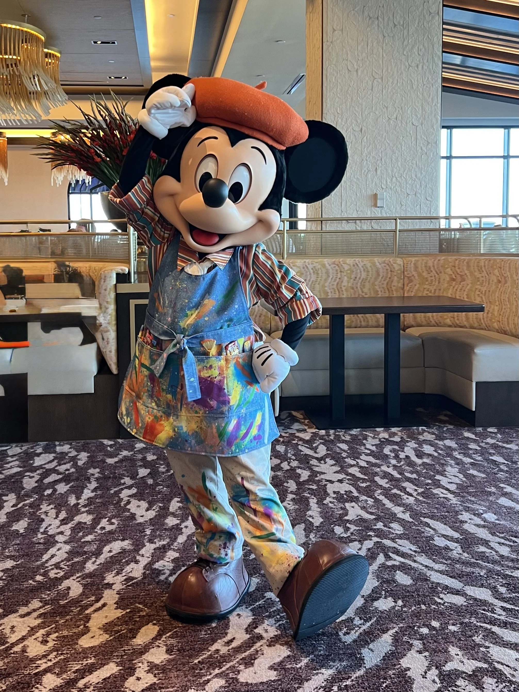 Mickey Mouse dressed as an artist, wearing a paint-splattered apron and pants, holding a beret, and standing on a patterned carpet in a restaurant or hotel lobby.