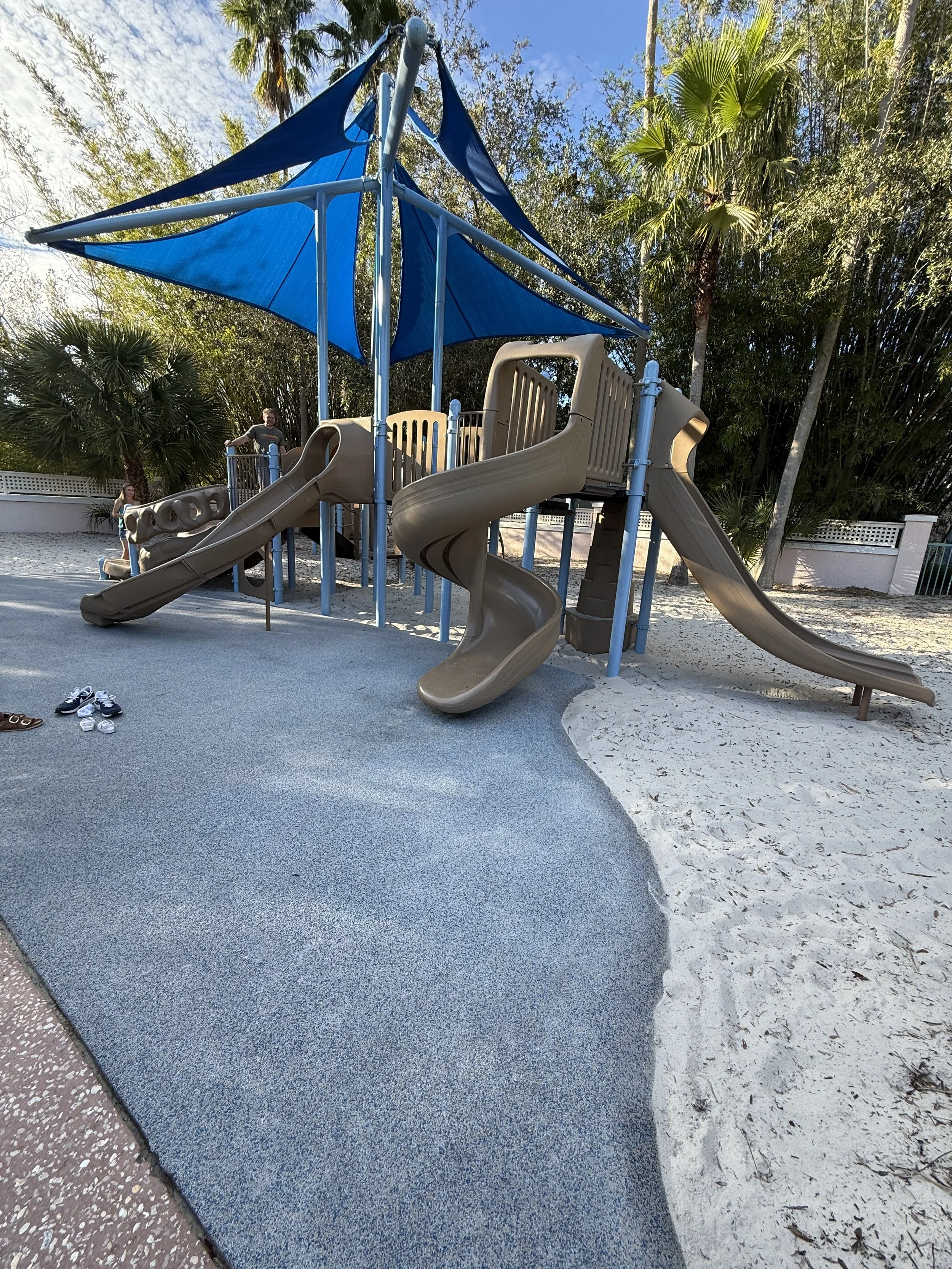 A playground with multiple slides, a small climbing structure, and a blue canopy, surrounded by trees and a sandy area.