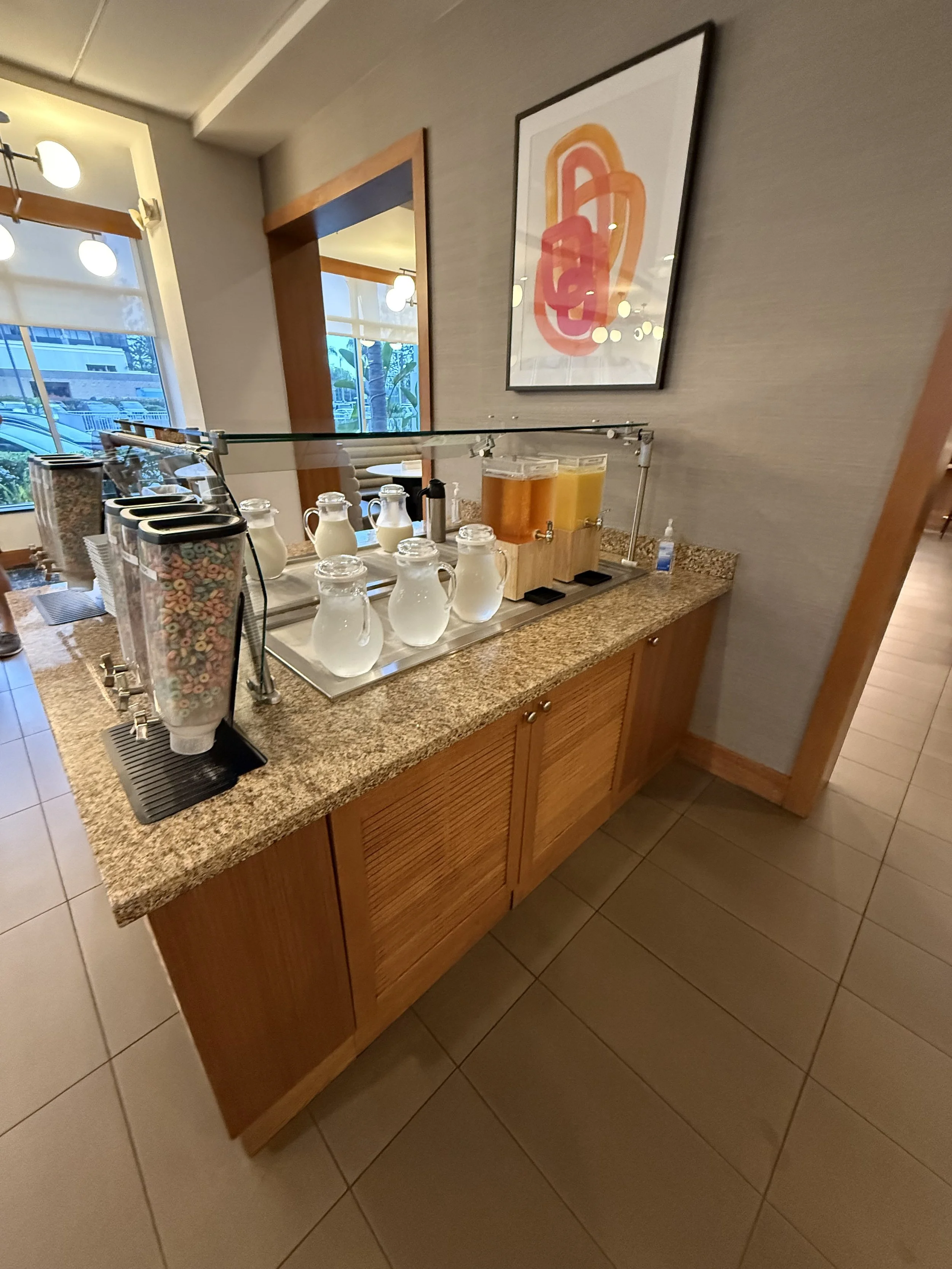 Breakfast juice and milk station with cereal dispenser and glass pitchers of milk and juice on a granite countertop in a restaurant.