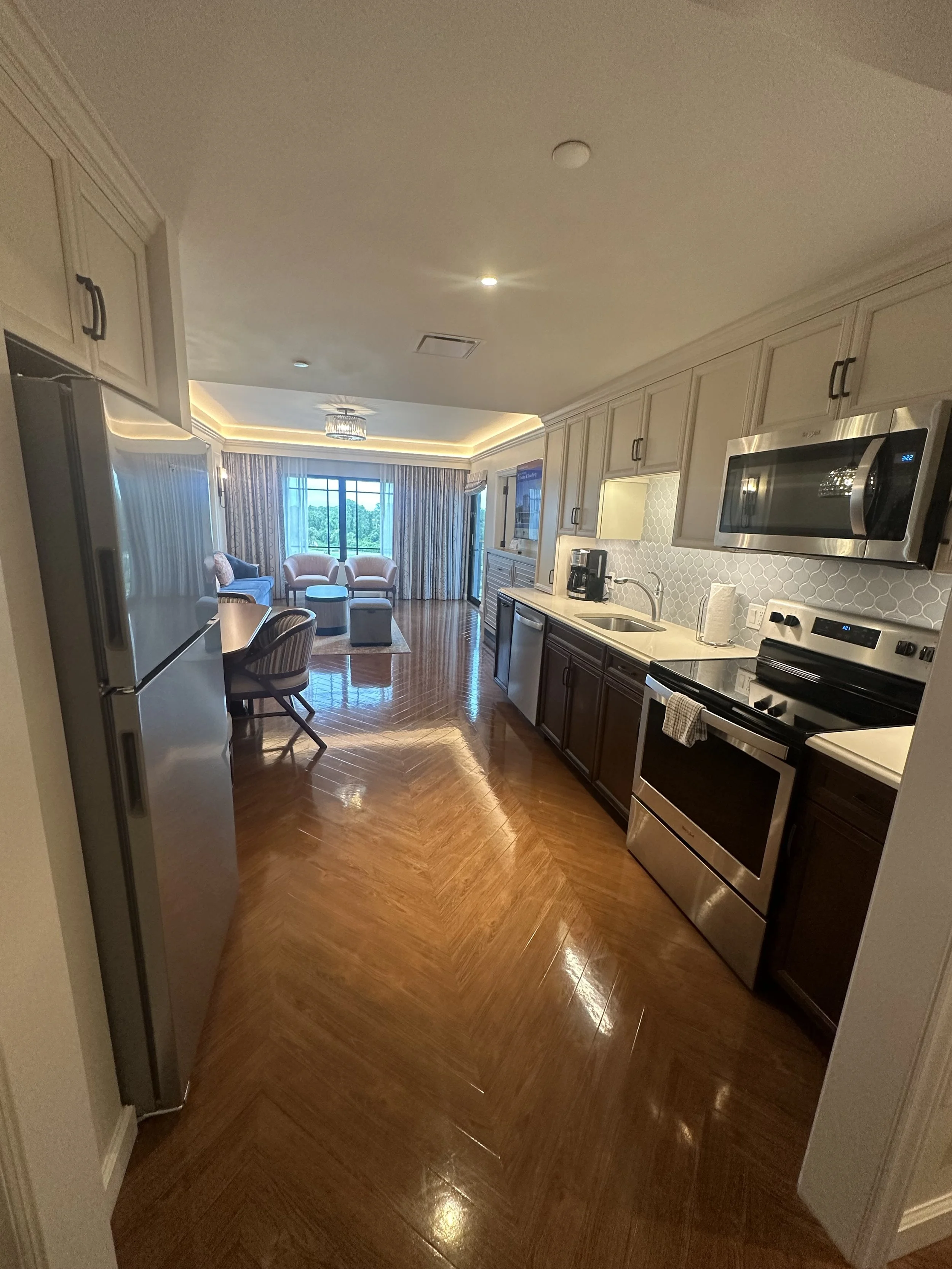 Modern kitchen with white and dark cabinets, stainless steel appliances, and a wooden herringbone floor, leading to a living room area with seating and large windows.