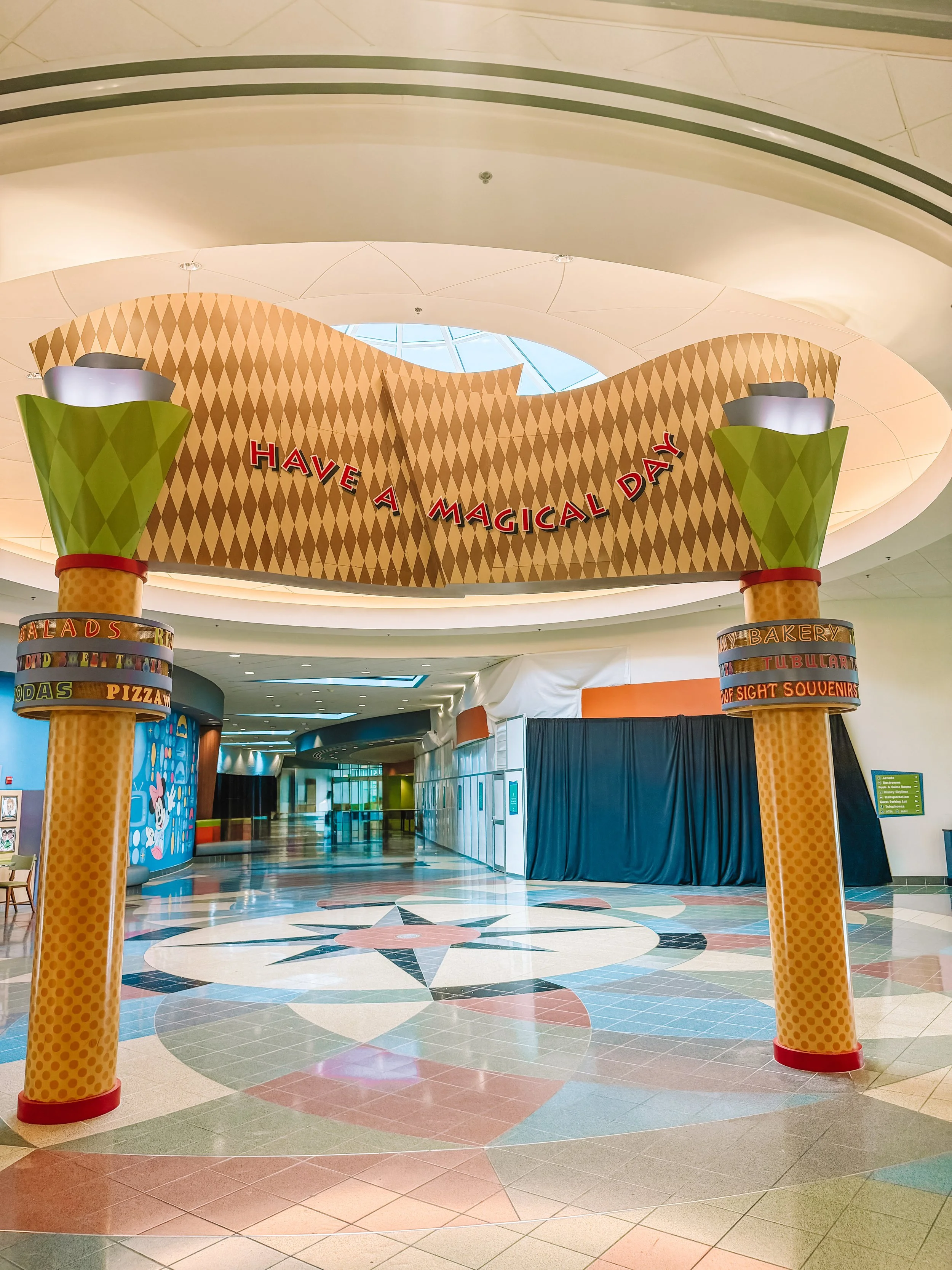 Decorative mall entrance arch with colorful columns and a sign that says, 'Have a Magical Day,' inside a shopping mall with a patterned tile floor.