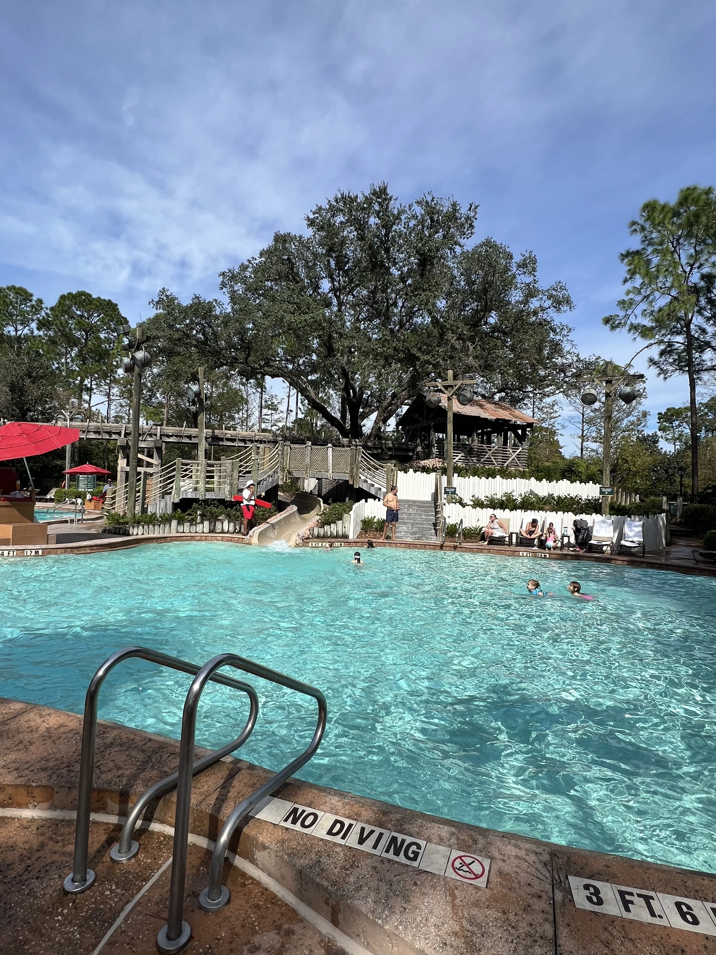 A swimming pool with people swimming and relaxing around the edges. There are trees and a wooden decked area with a hut in the background. A sign on the pool edge reads 'No Diving' and indicates a depth of 3 feet 6 inches.