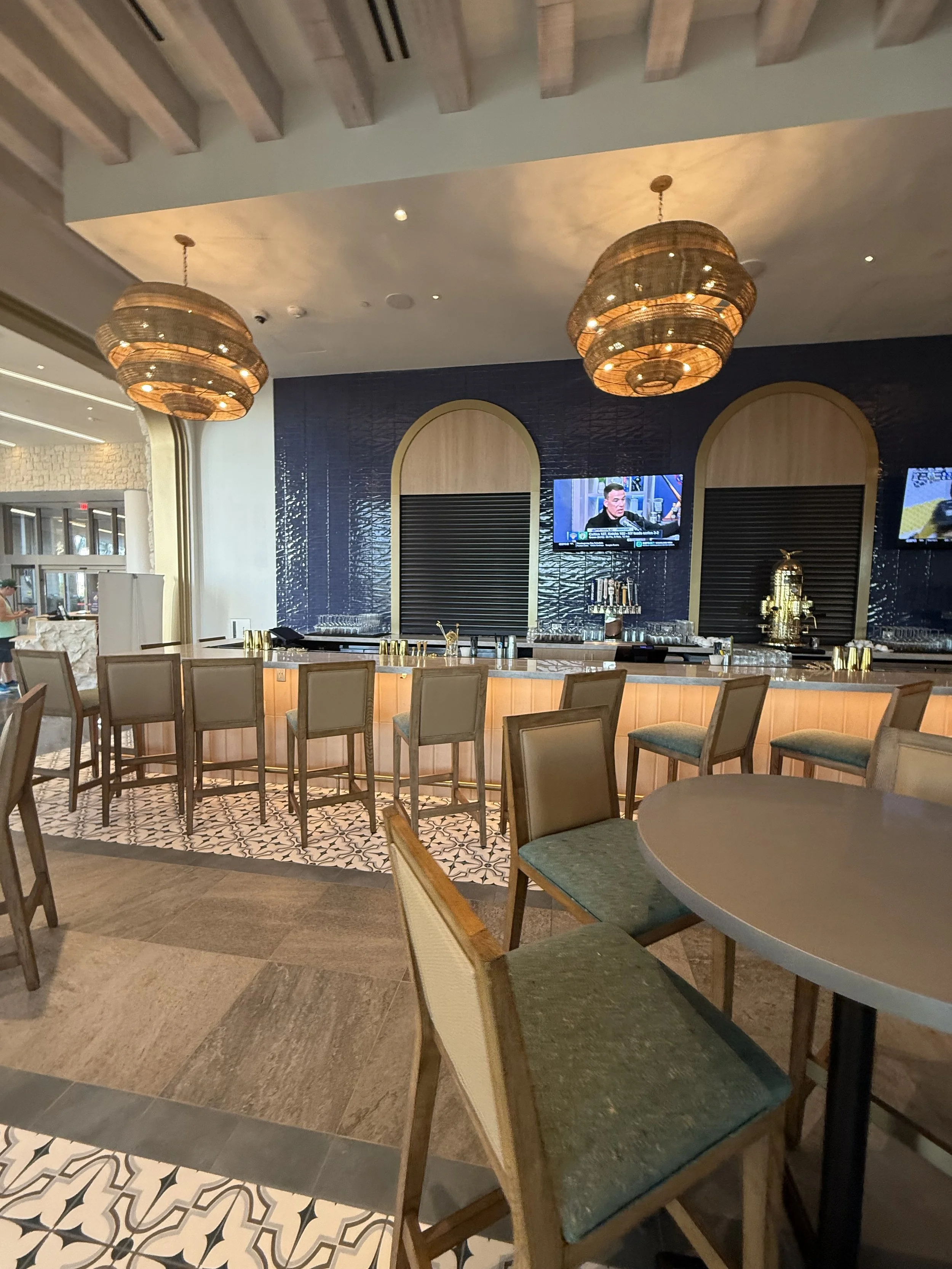 Interior of a modern bar with high chairs, a illuminated bar counter, and decorative hanging light fixtures. Two television screens on a navy blue wall display news programs.