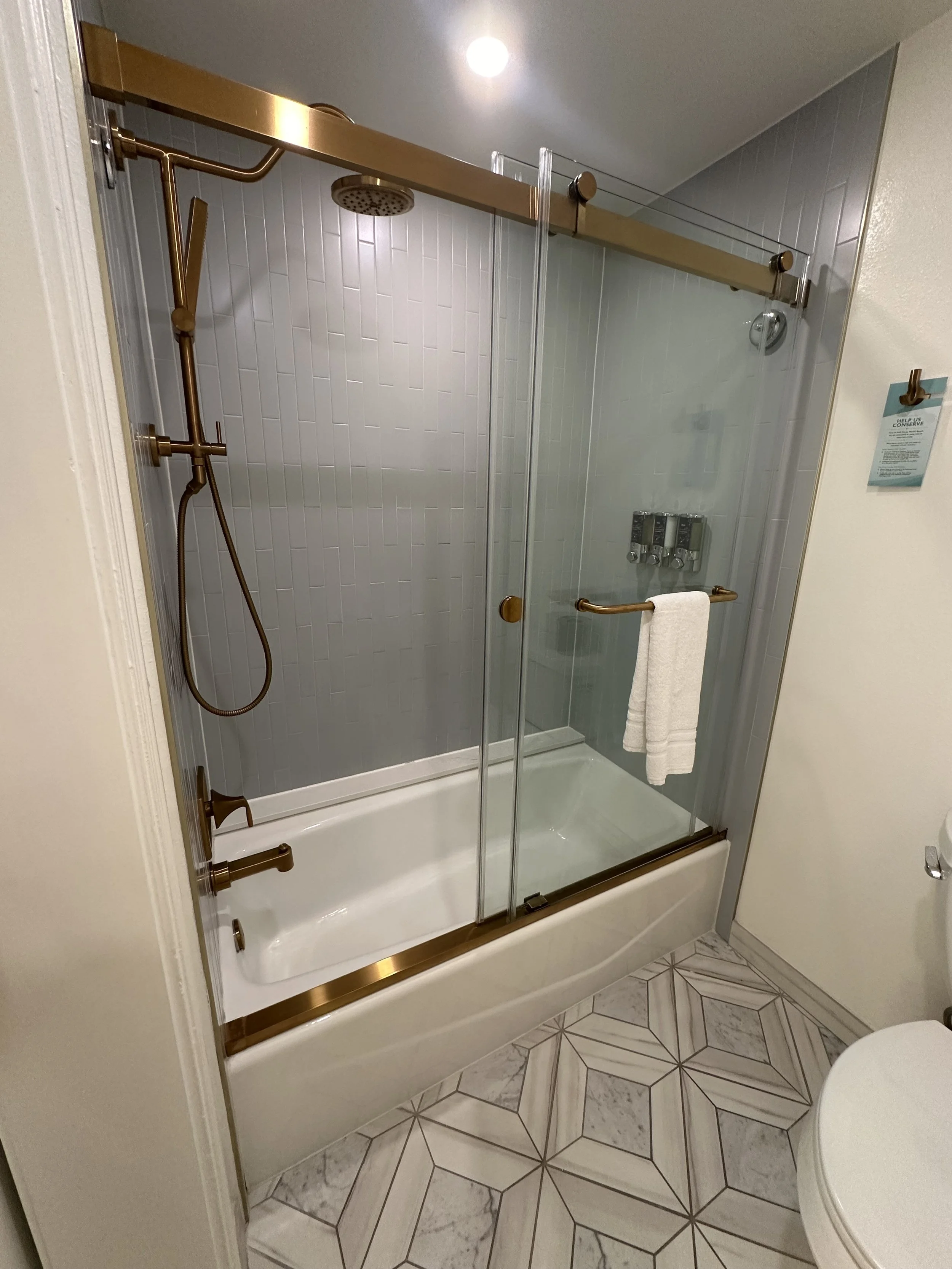 Bathroom shower with glass sliding doors, gold fixtures, a bathtub, a rain shower head, and a handheld shower head. There is a white towel hanging on the door handle.
