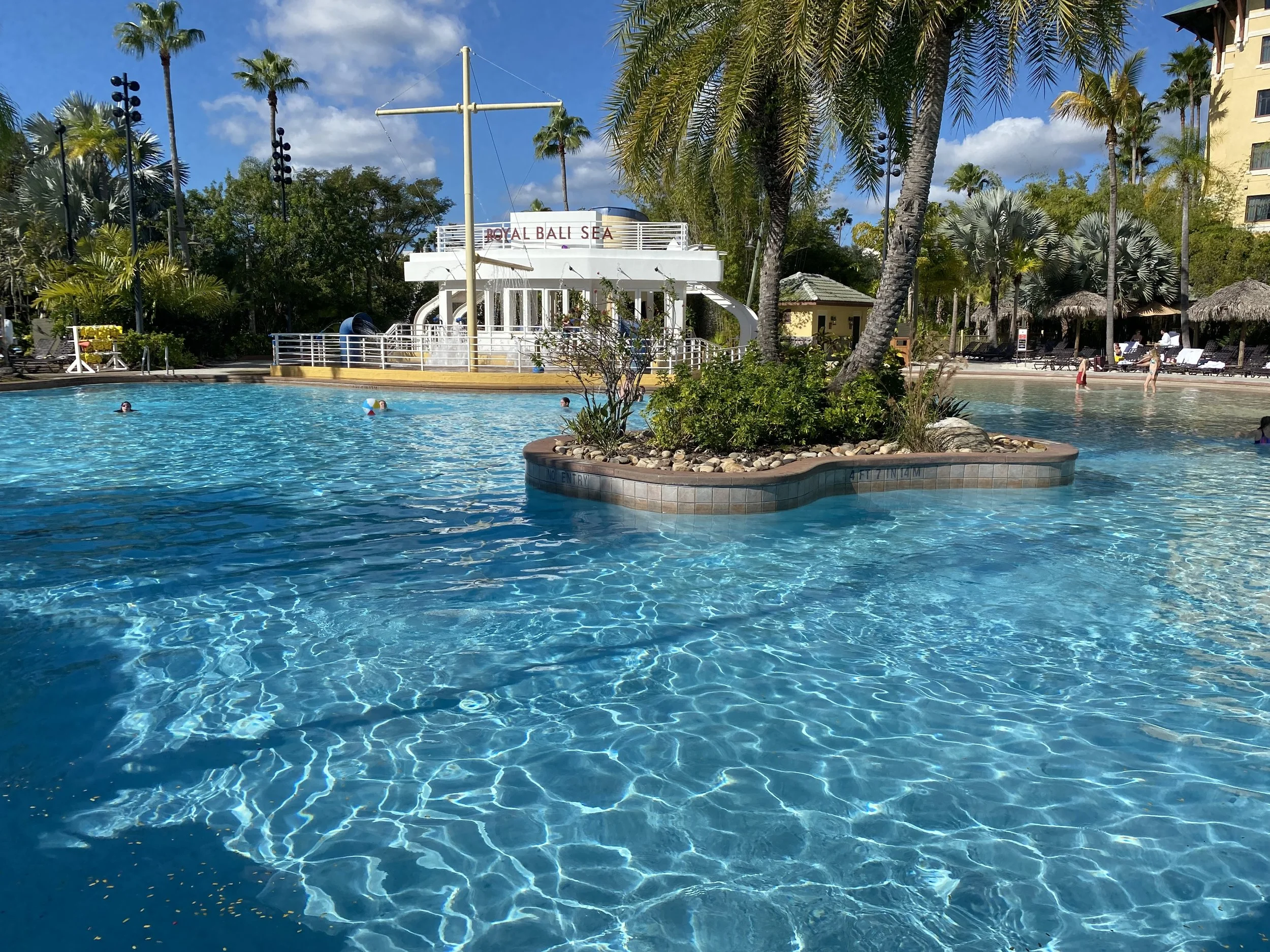Swimming pool with a small island of rocks and palm trees in the water, surrounded by lounge chairs and umbrellas, with a white boat-shaped structure labeled 'Royal Bali Sea' in the background, and people swimming and relaxing under a partly cloudy s