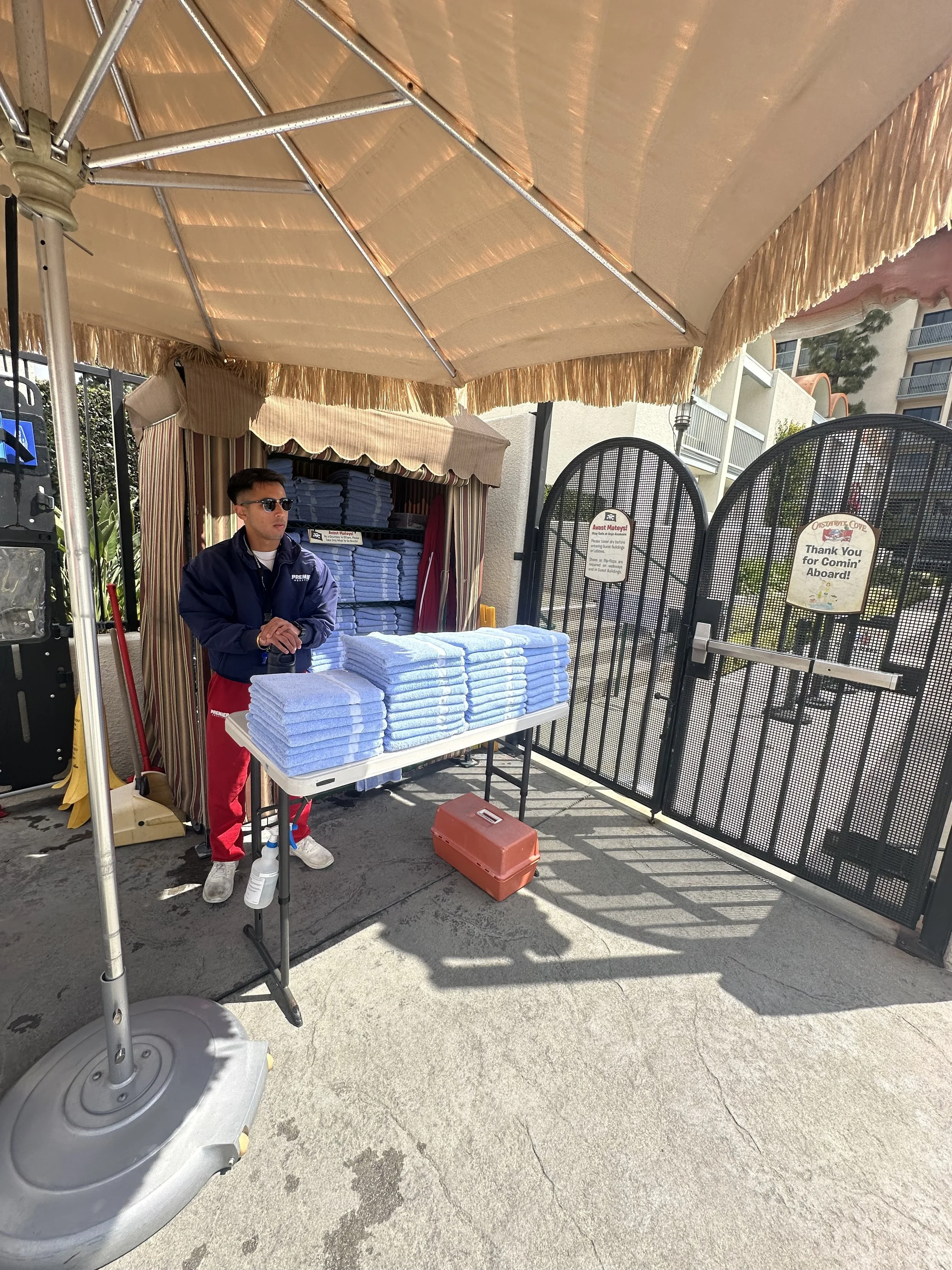 A man wearing sunglasses and a blue jacket standing behind a table with neatly folded blue towels under a large beige canopy at an outdoor entrance gate to a pool or beach area, with open gate and signs on the gate.