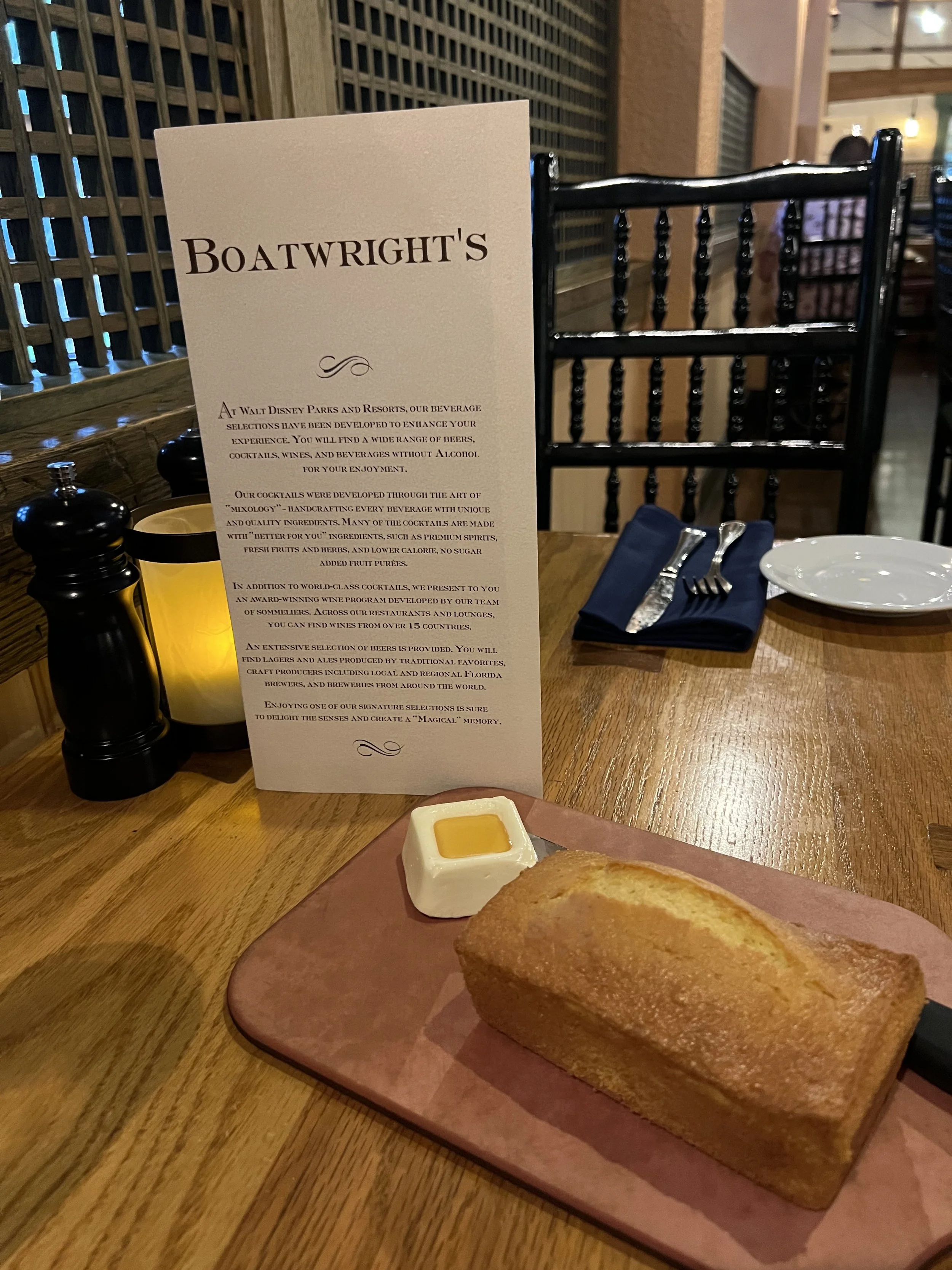 A table set with a pink plate holding a loaf of bread and a small dish of butter, a blue cloth napkin with silverware, a white plate, a candle, pepper and salt shakers, and a menu card describing beverages at Walt Disney Parks and Resorts.