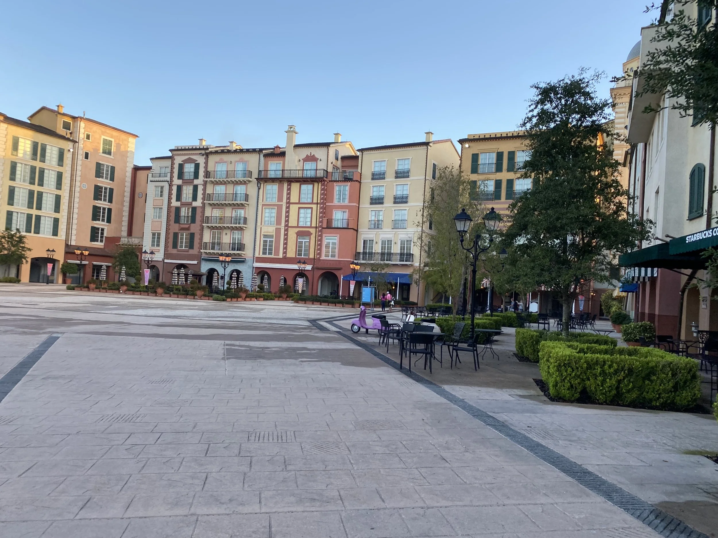 Empty plaza with colorful European-style buildings, outdoor seating, benches, street lamps, trees, and a Starbucks.