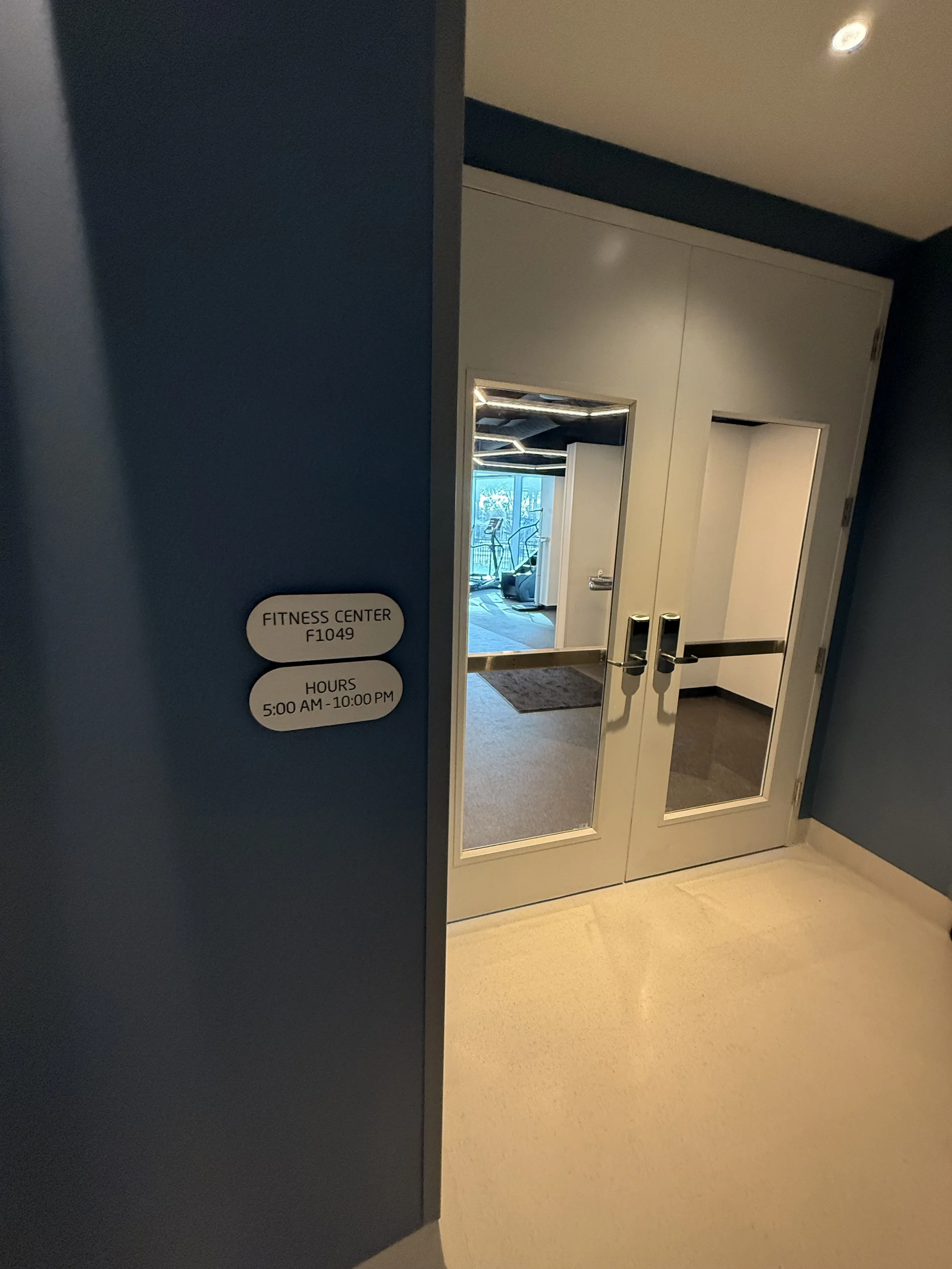 Entrance to a fitness center with blue walls and double glass doors. A sign indicates the fitness center is room F1049, open from 5:00 AM to 10:00 PM. Inside, exercise equipment is visible.