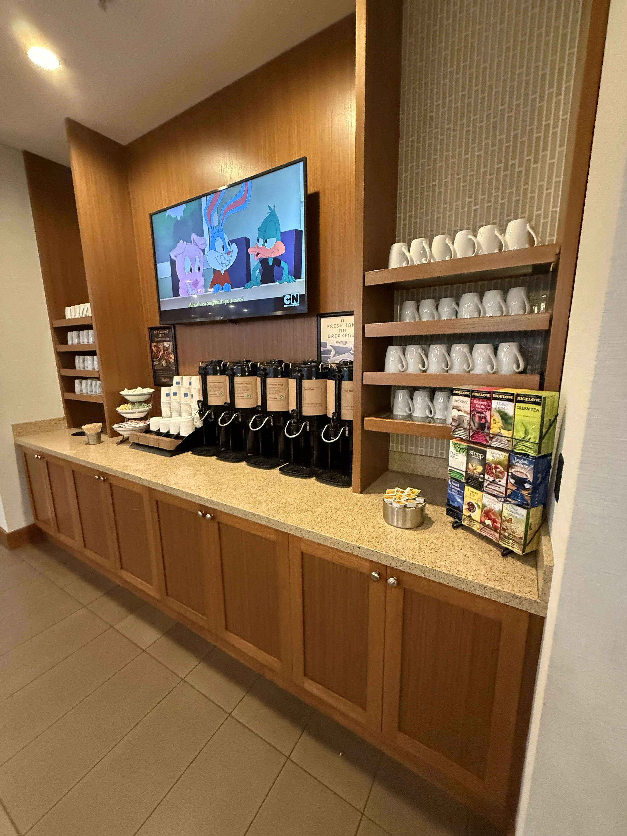 A breakfast coffee station with coffee dispensers, cups, tea bags, and a tv screen showing cartoon characters.