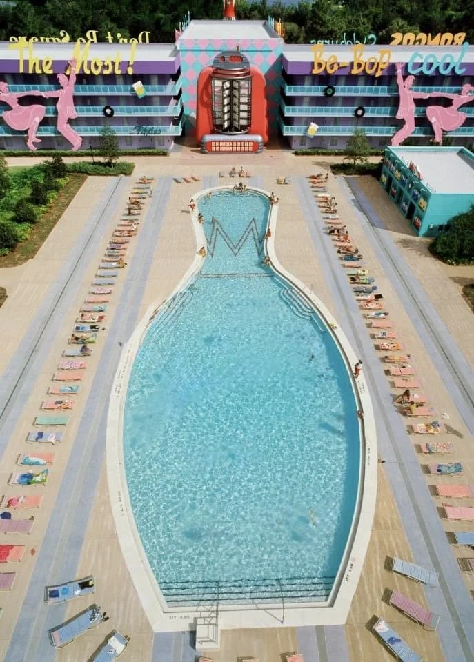 An aerial view of a large swimming pool with beach chairs along its sides, in front of a colorful building with a bright red, cartoon-style apparent vending machine design on the front. The building has large pink rabbit and yellow slime figures, wit