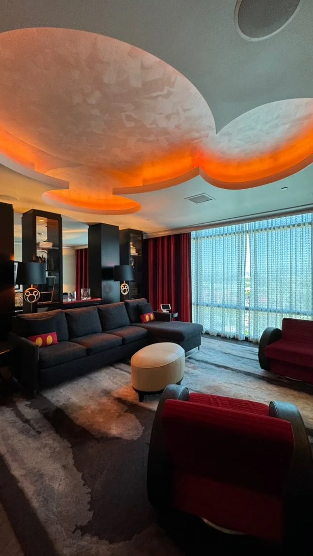 Modern living room with a dark sofa, red armchair, and beige ottoman, large window with sheer curtains, illuminated ceiling feature, and a patterned rug.