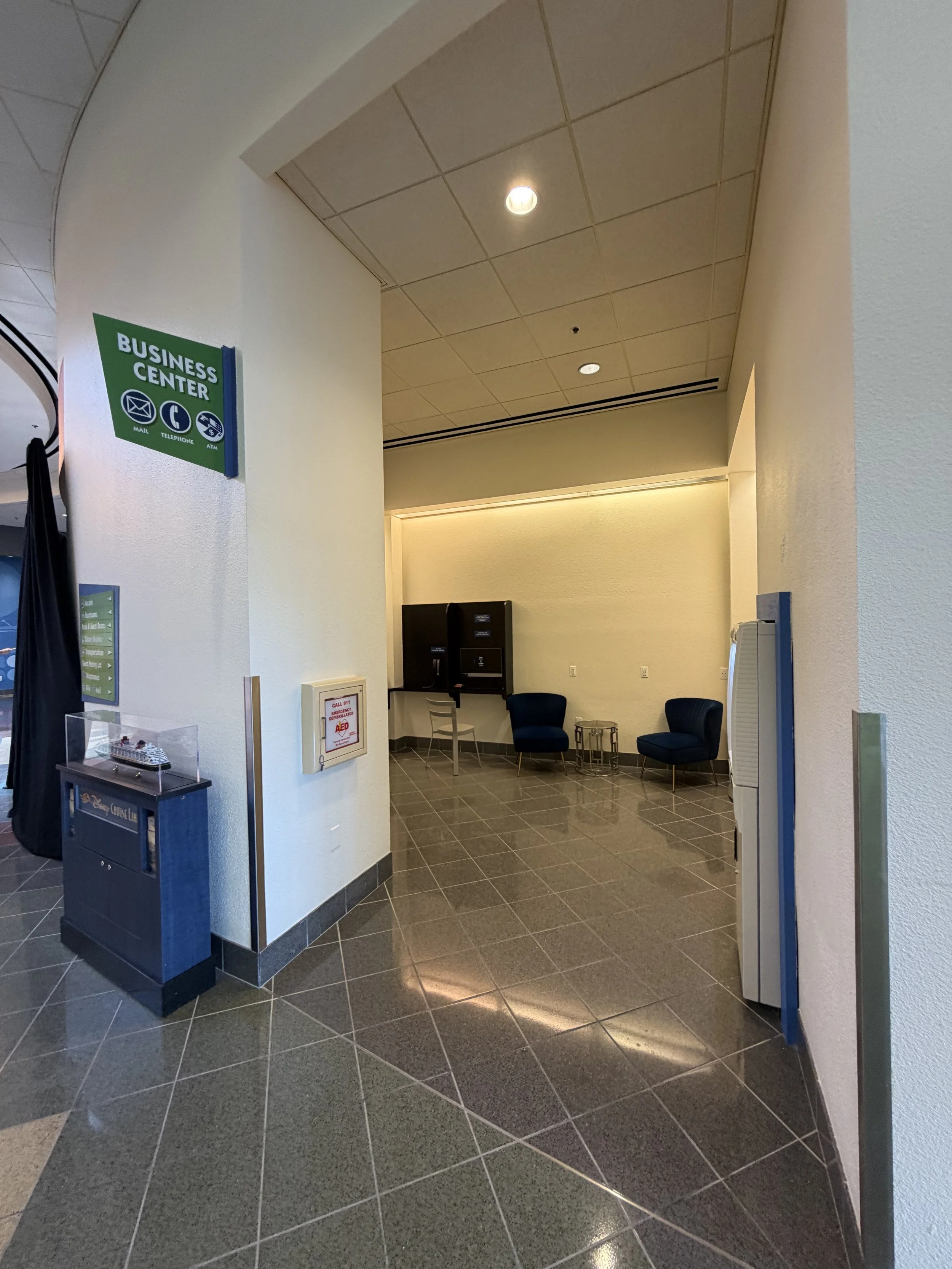 Interior of a business center waiting area with two blue chairs, a glass table, and an ATM or kiosk, with a wall in the background.