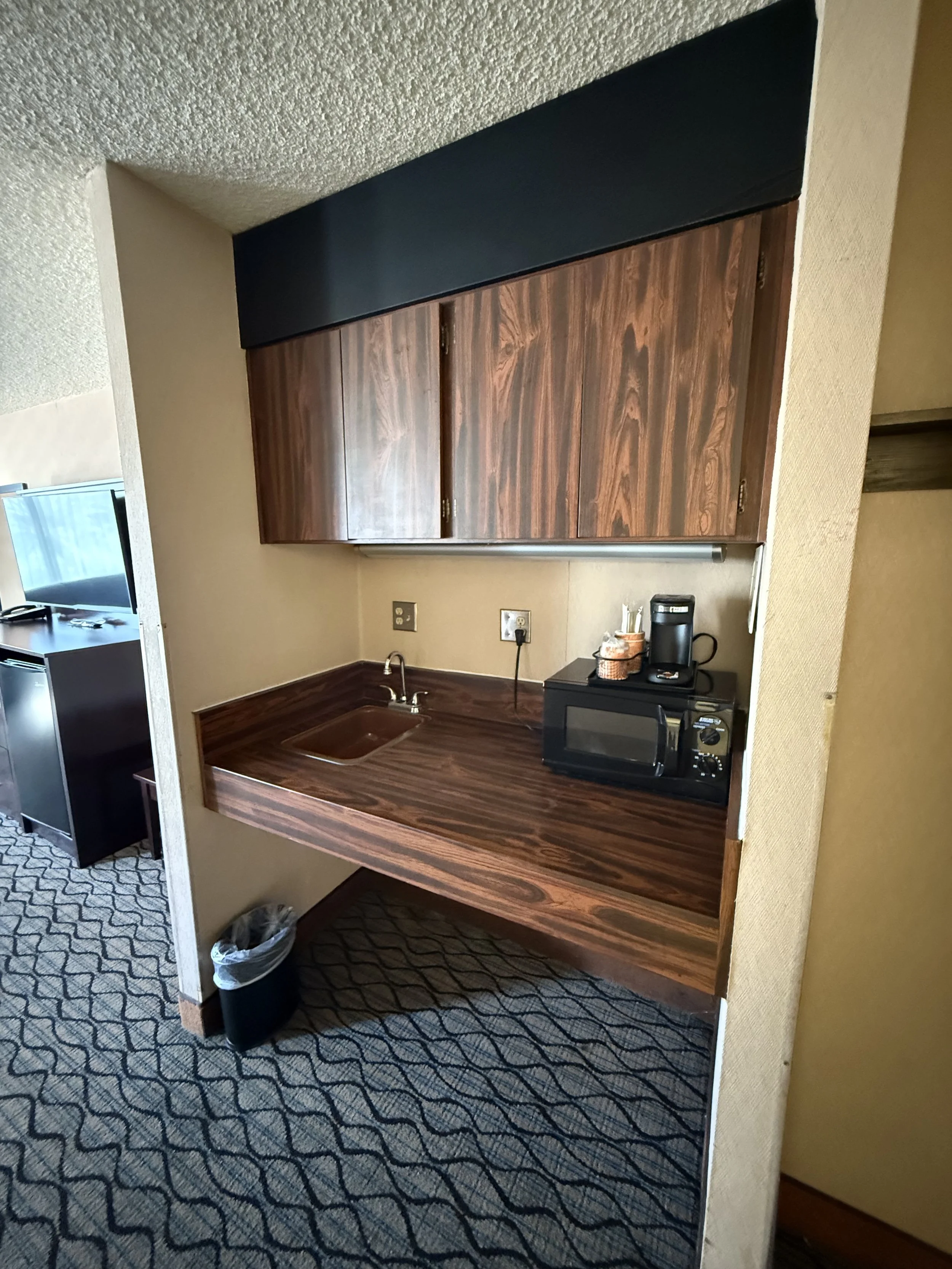 Compact kitchenette with wooden cabinets, a small sink, microwave, coffee maker, and trash can, in a room with patterned carpet.