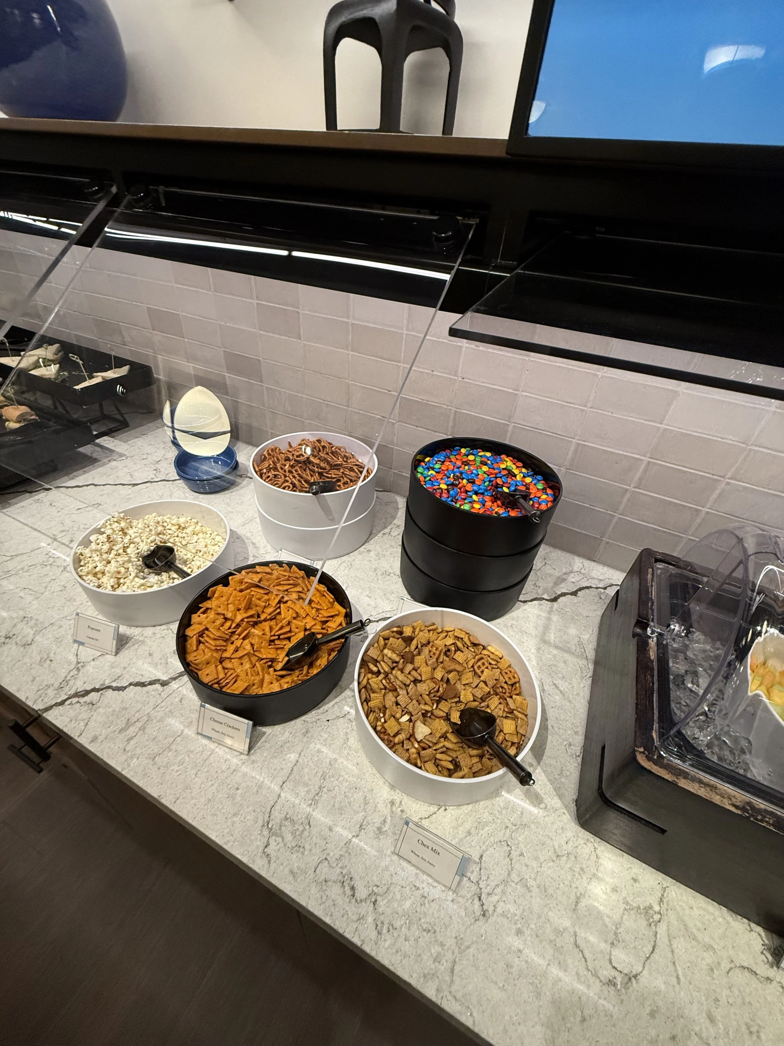 A cereal breakfast bar with bowls of popcorn, chocolate cereal, spicy snack mix, and other cereals on a granite countertop.