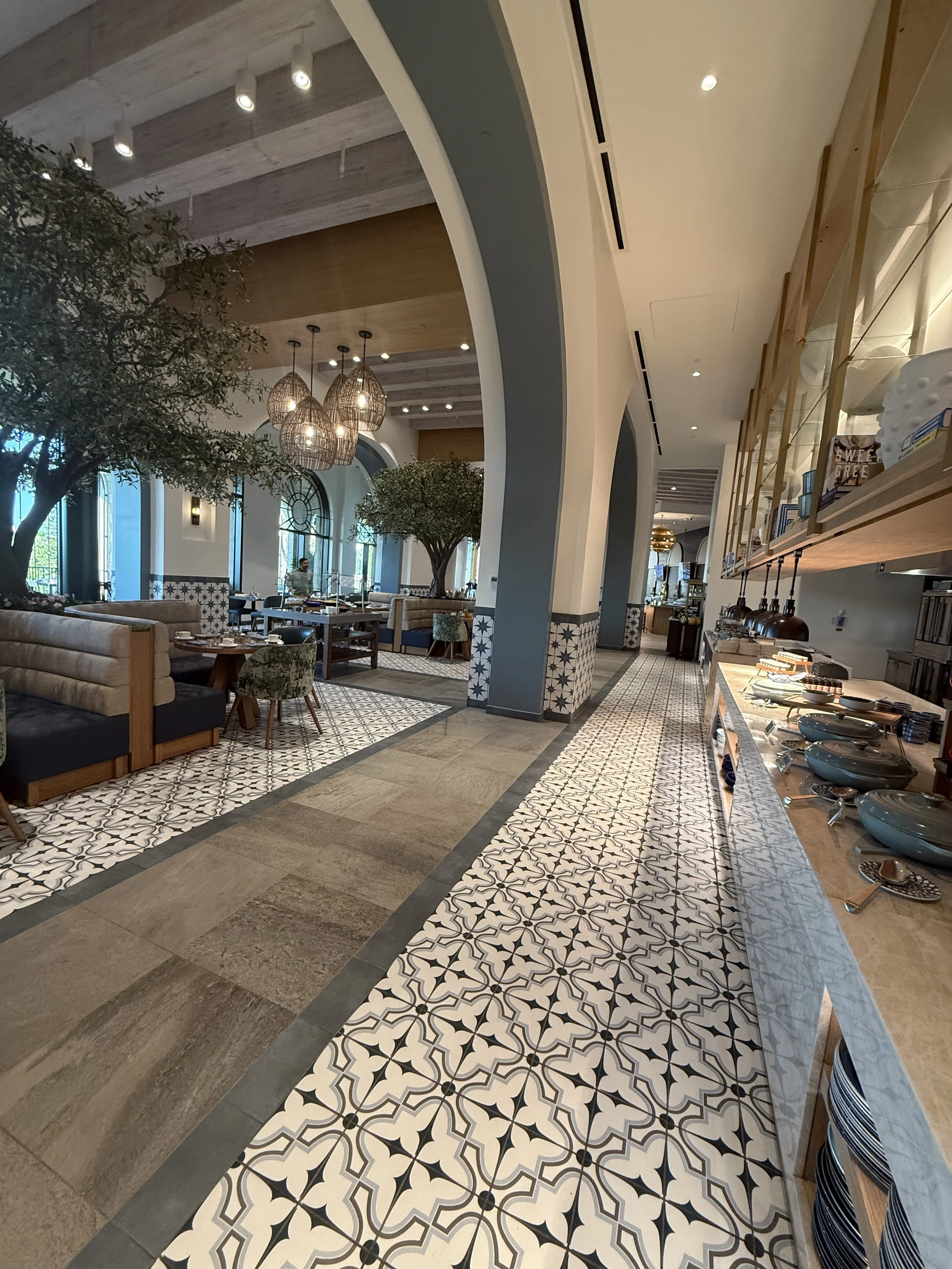 Interior of a restaurant with patterned tile flooring, arched doorways, dining tables and chairs, greenery, and hanging light fixtures.