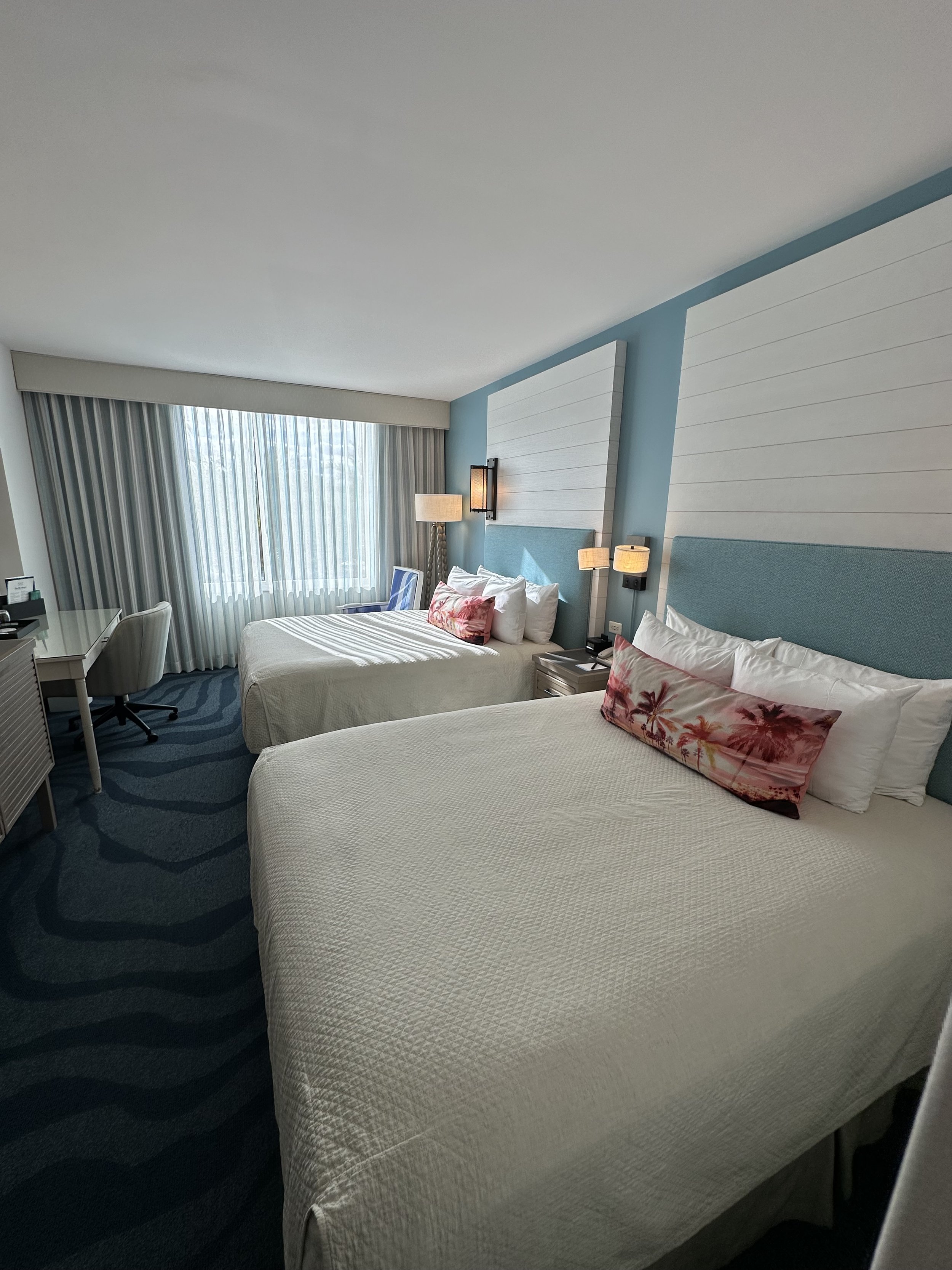 Hotel room with two queen beds, blue headboards, pink palm tree pillows, nightstands, lamps, a desk with a chair, and a large window with curtains.