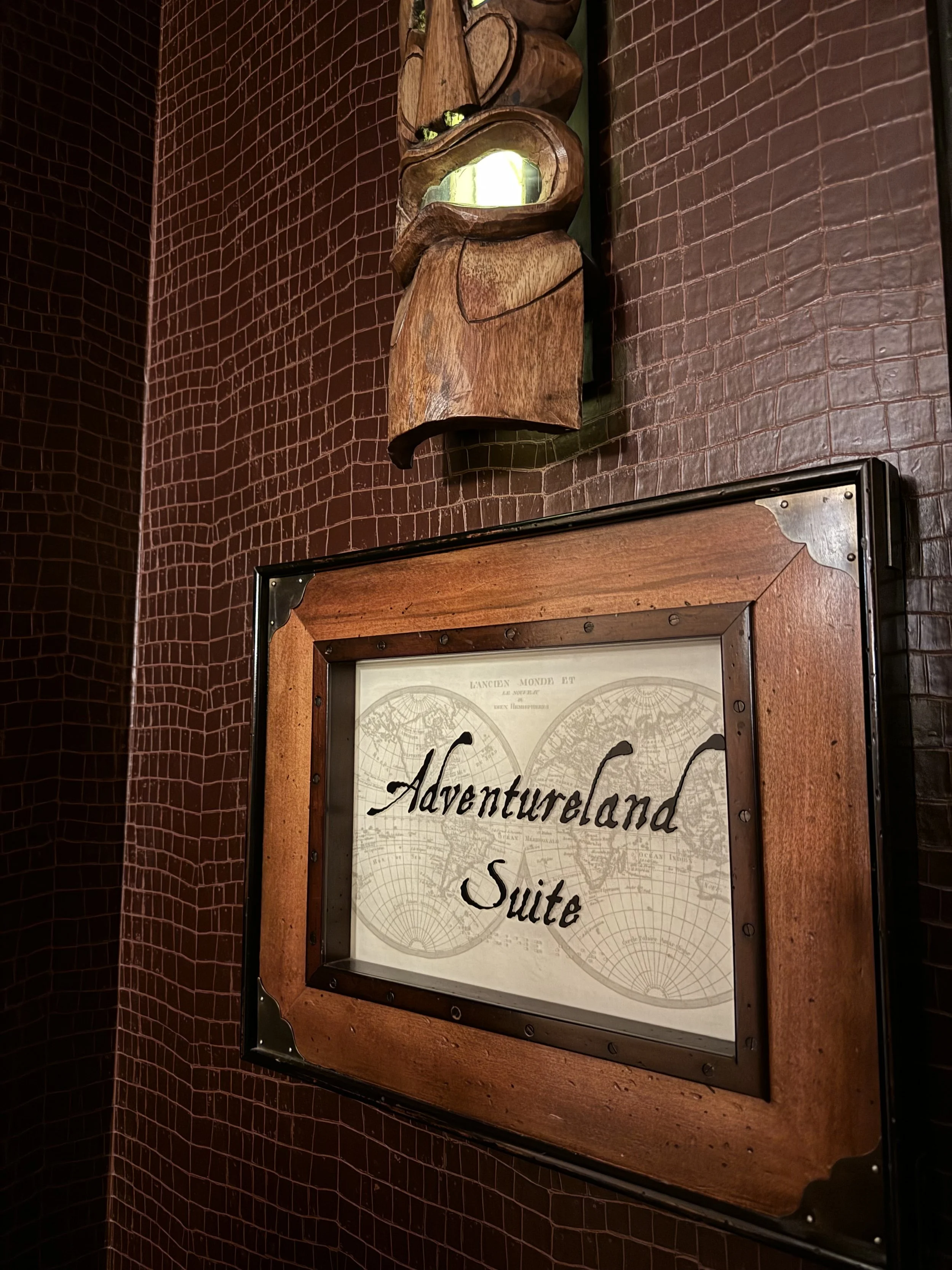 Sign for Adventureland Suite in a wooden frame on a red brick wall. Above it, a wooden sculpture or decorative piece is mounted on the wall.