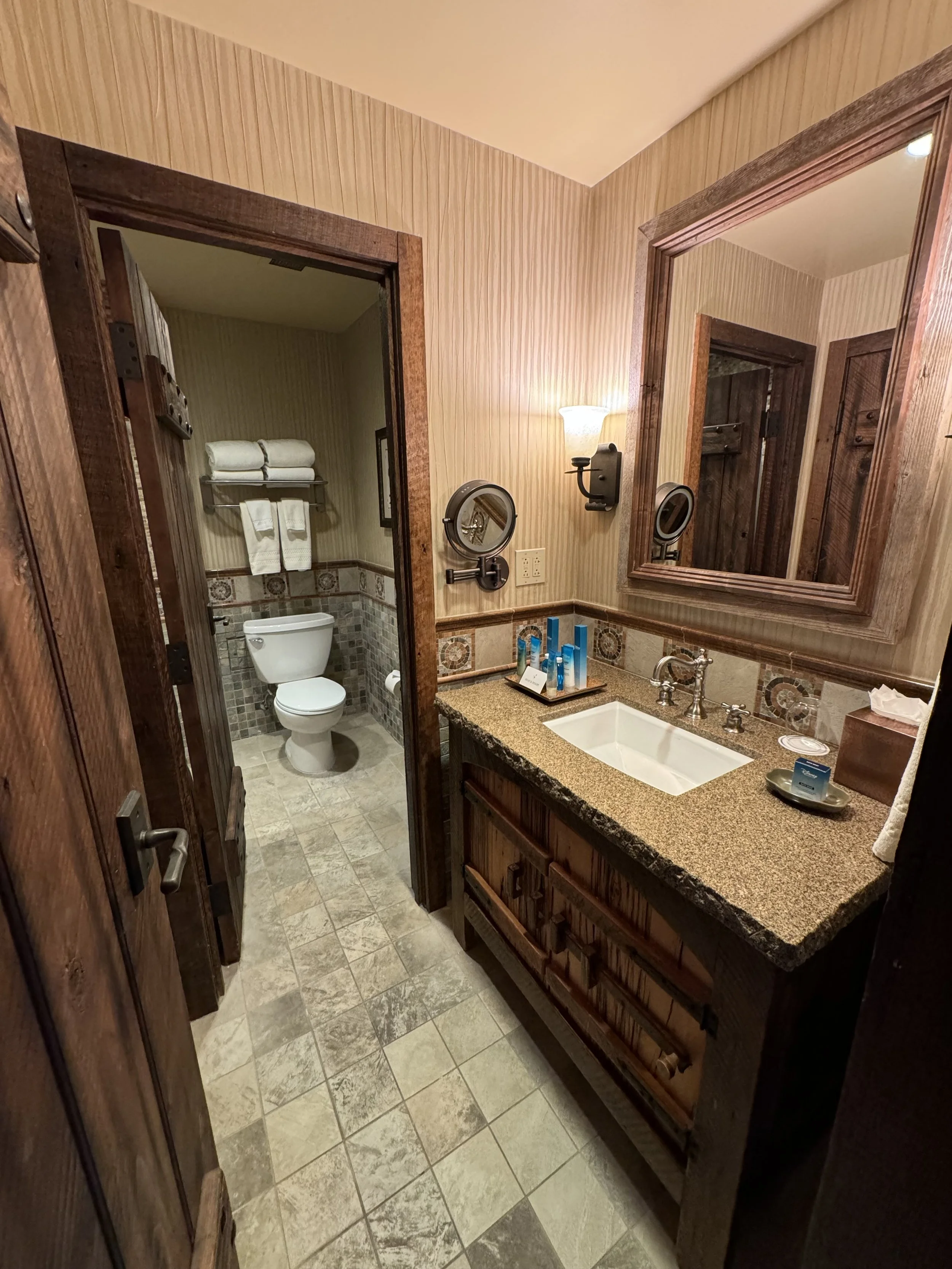 A rustic bathroom with wood-paneled walls, a granite countertop vanity with a white sink, a large mirror, wall-mounted light fixture, and a mirror with a built-in magnifying mirror. In the adjacent room, a toilet with towels on a shelf above is visib