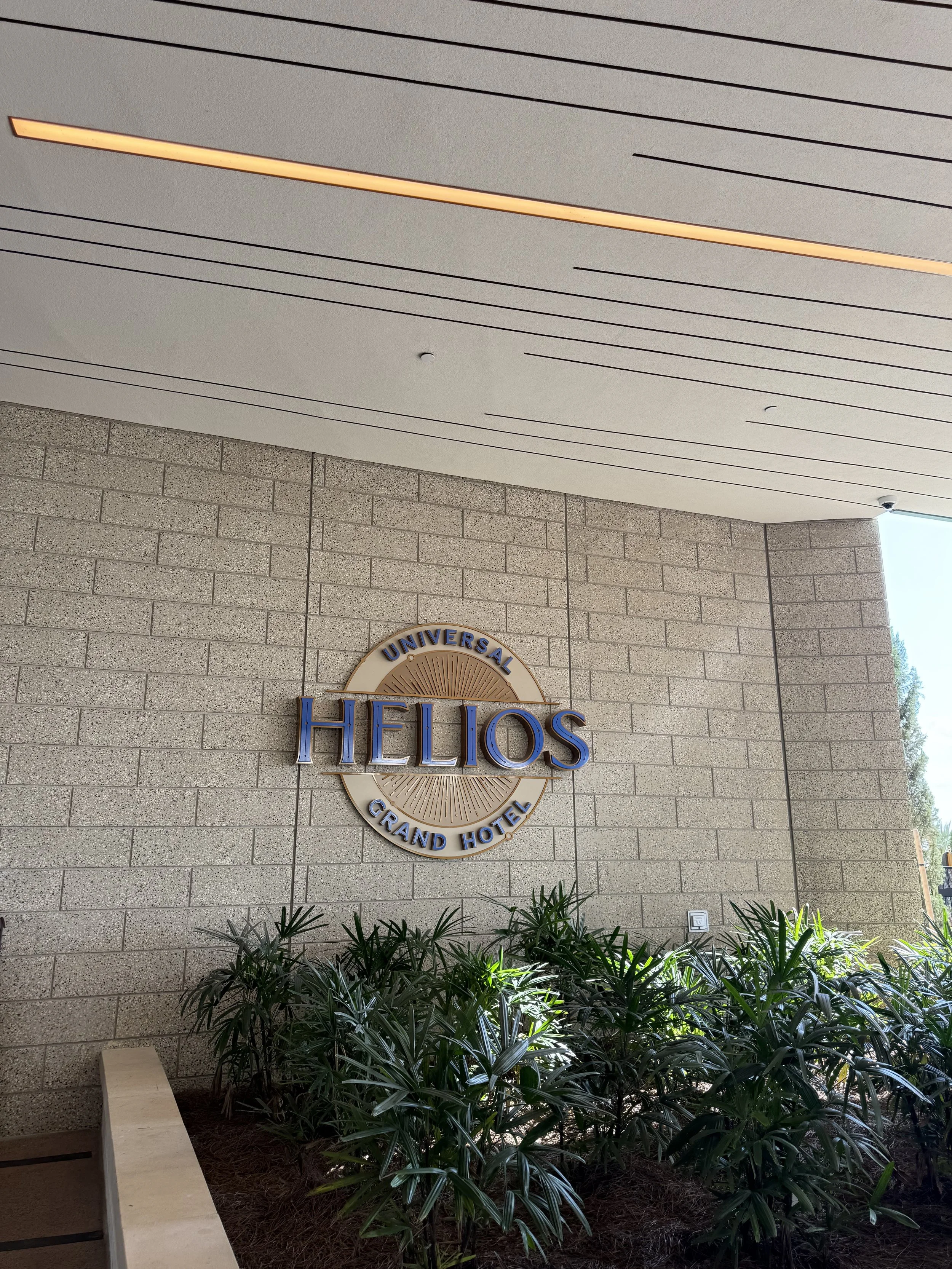 Sign for Universal Helios Grand Hotel on a beige brick wall, with green plants in front.