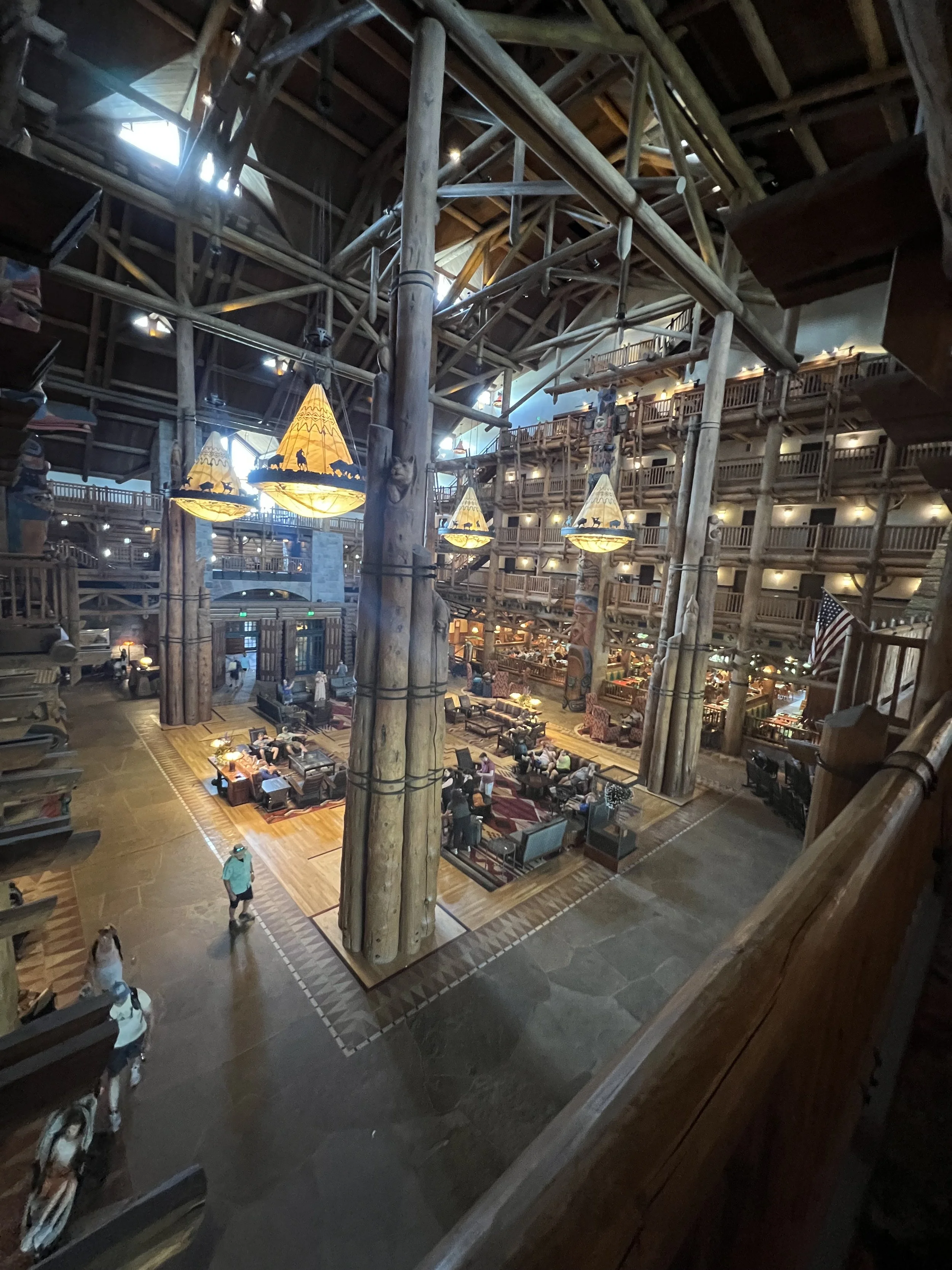 Interior of a large, multi-story lodge with wooden logs and beams, featuring a spacious central area with seating and decorative hanging lamps, and balconies overlooking the main hall.
