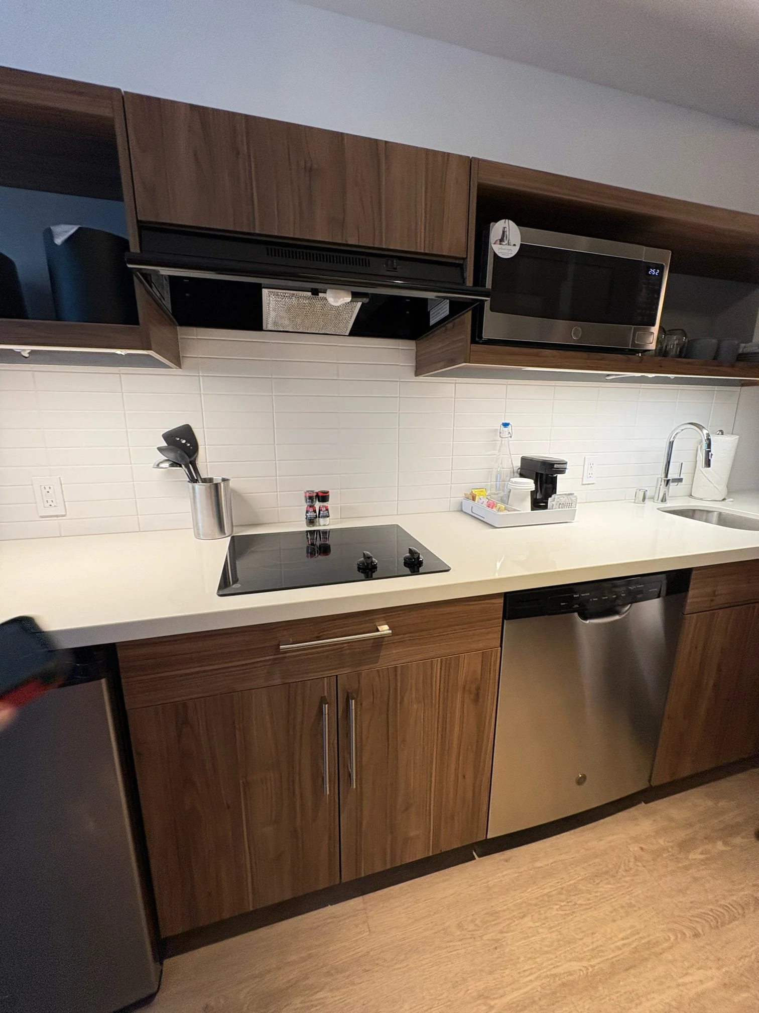 Modern kitchen with wooden cabinets, white countertop, black stovetop, microwave, and stainless steel dishwasher. Counter has a small tray with beverages, a water bottle, and coffee supplies.