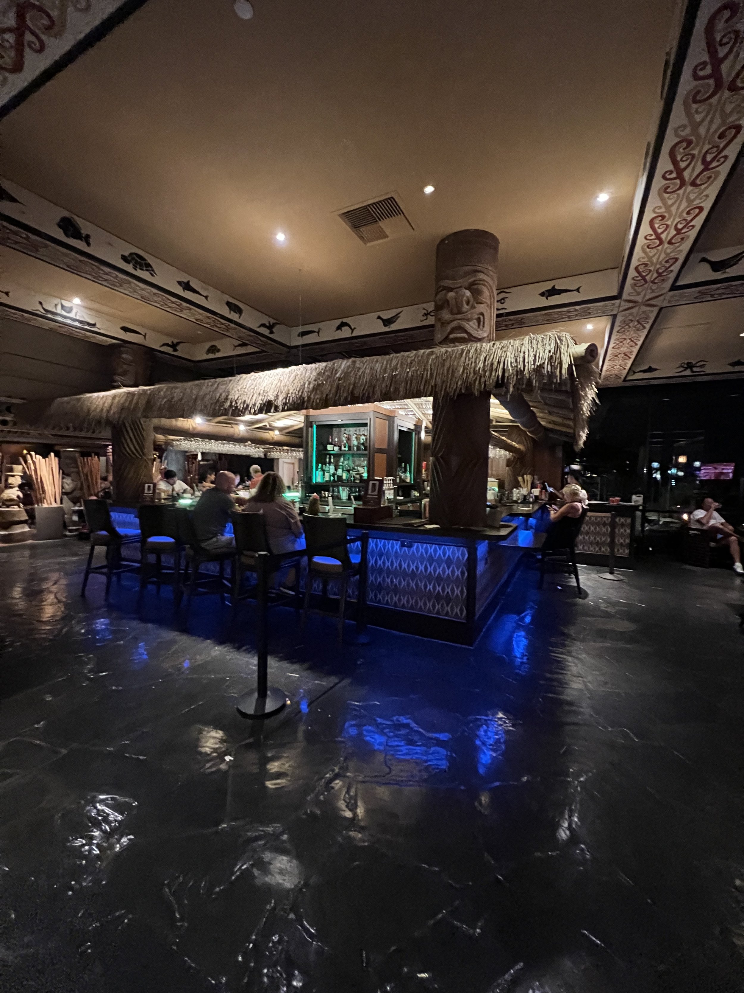 Indoor bar with tiki theme, featuring tiki statues and thatched roof decoration, with a few patrons sitting at the bar and socializing.