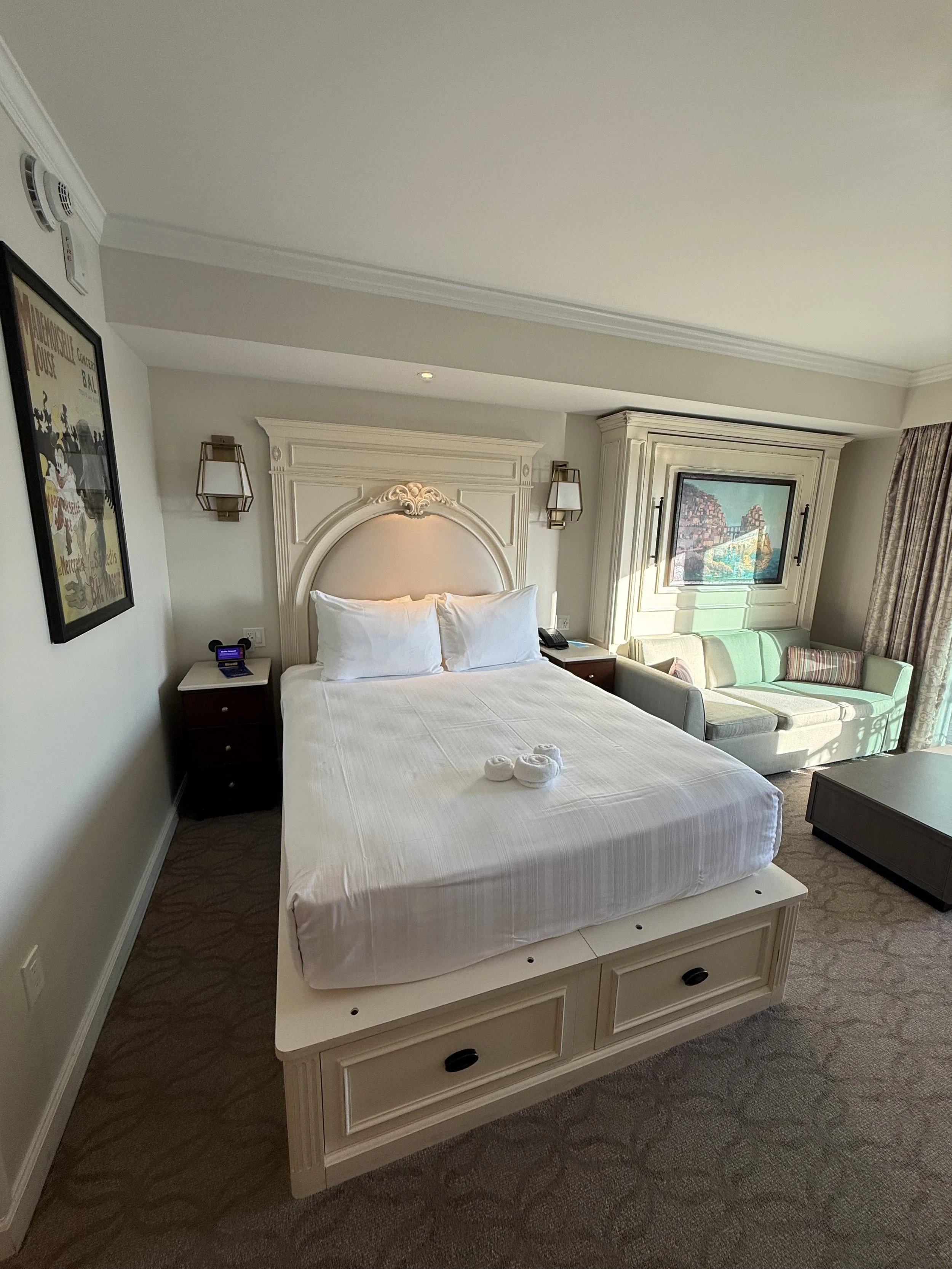 Hotel room with a neatly made bed, a sofa with cushions, a wall picture, and a window with curtains.