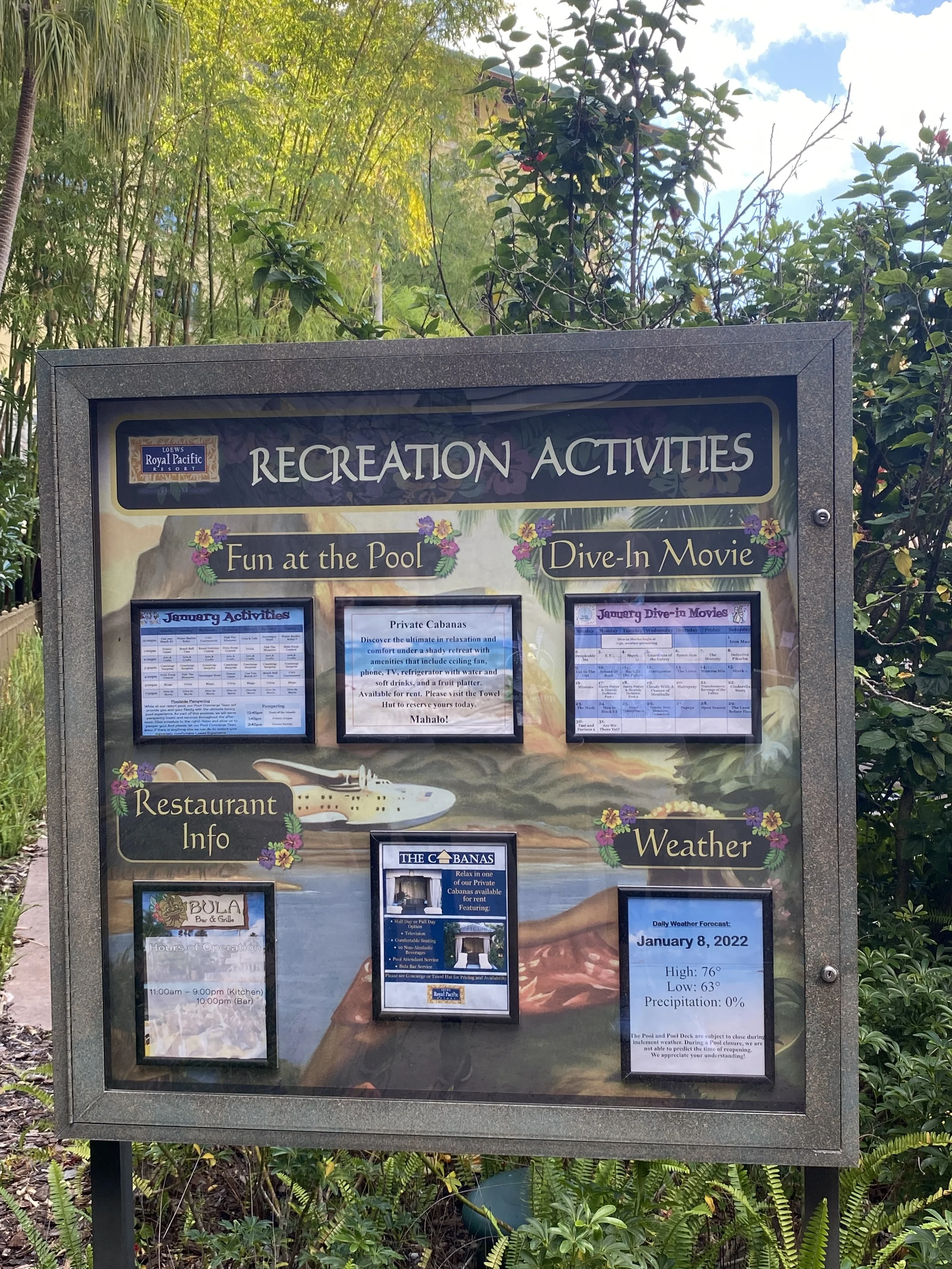 Recreation activities board at a resort displaying information about fun at the pool, dive-in movies, restaurant hours, weather forecast, and private cabanas, surrounded by green foliage and trees.