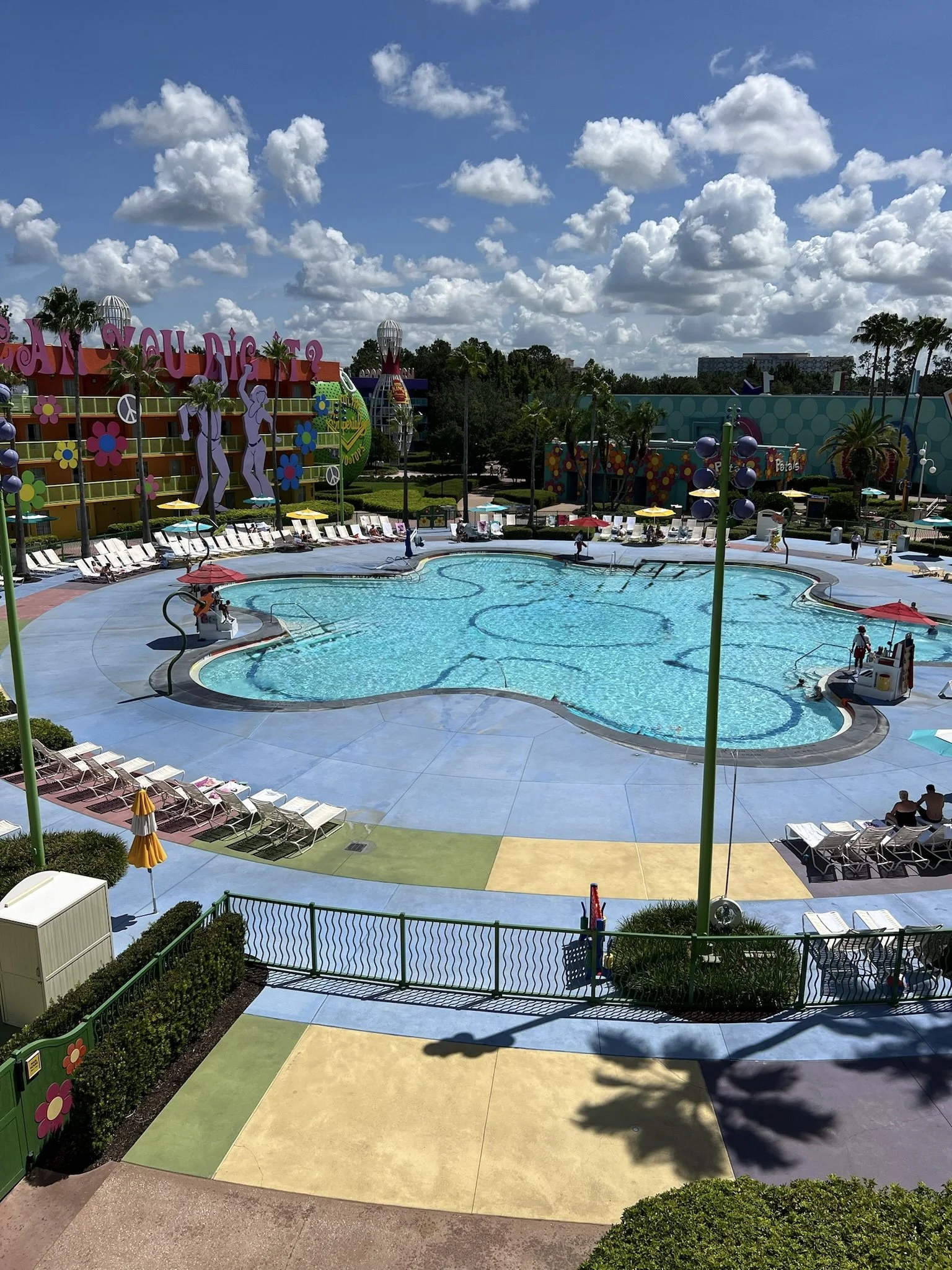 Empty colorful swimming pool with lounge chairs and umbrellas, surrounded by a playful, vibrant amusement park setting with palm trees and cartoon-like decorations.