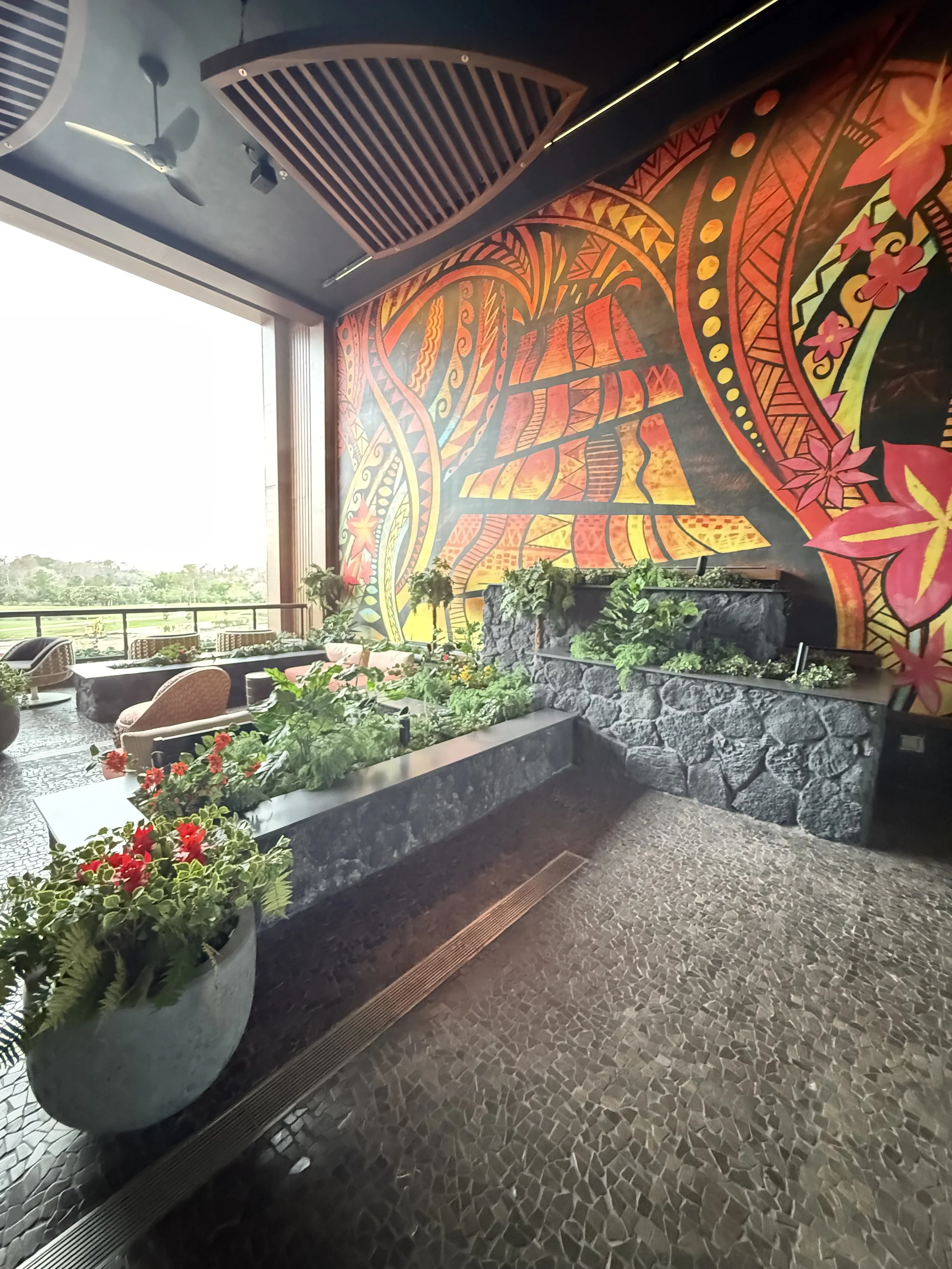 Indoor lounge area with a large colorful mural on the wall, potted plants, and outdoor scenery visible through large windows.