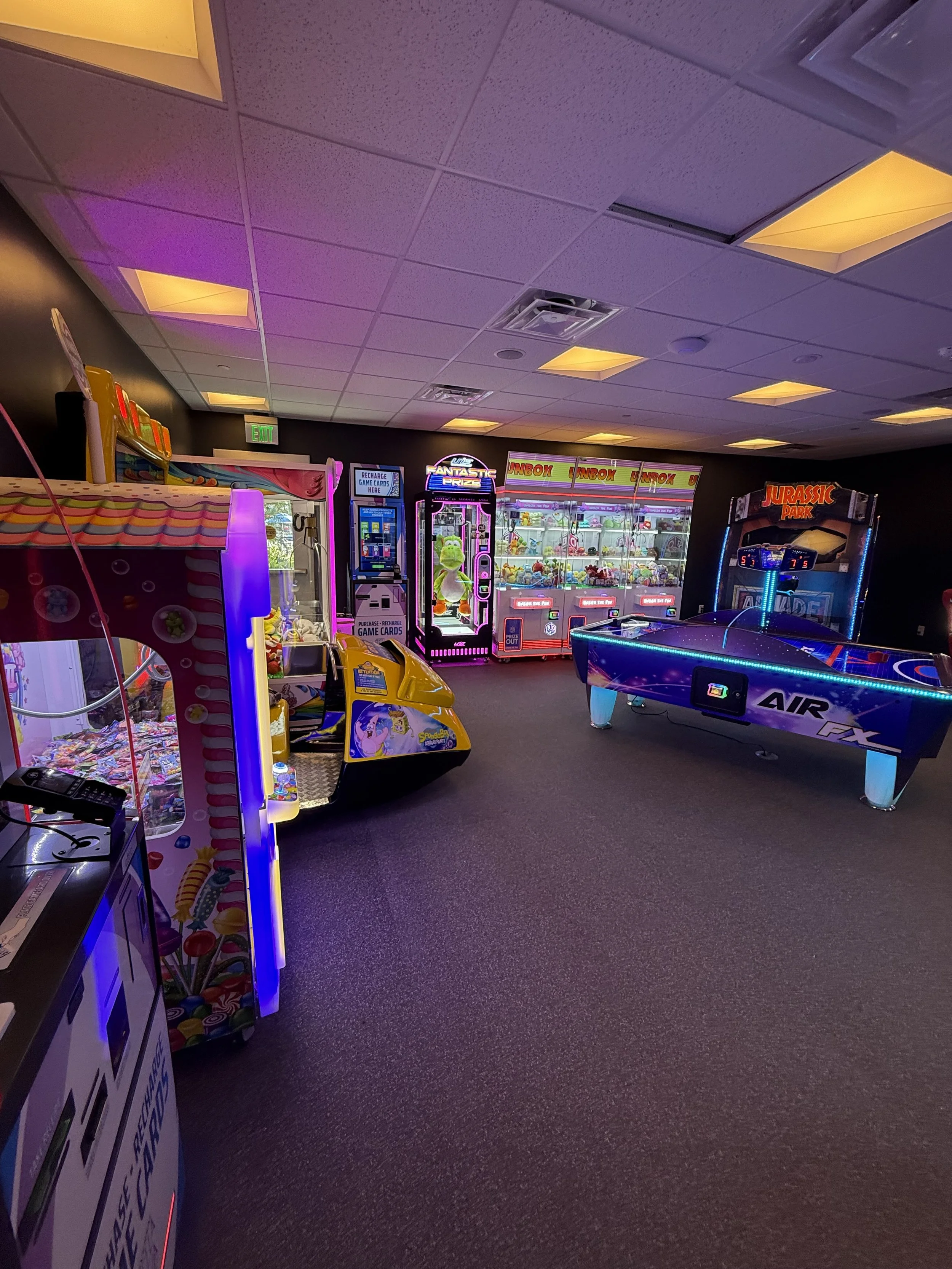 An indoor arcade room with various game machines including claw machines, a skee-ball, and air hockey tables, illuminated by colorful lights.