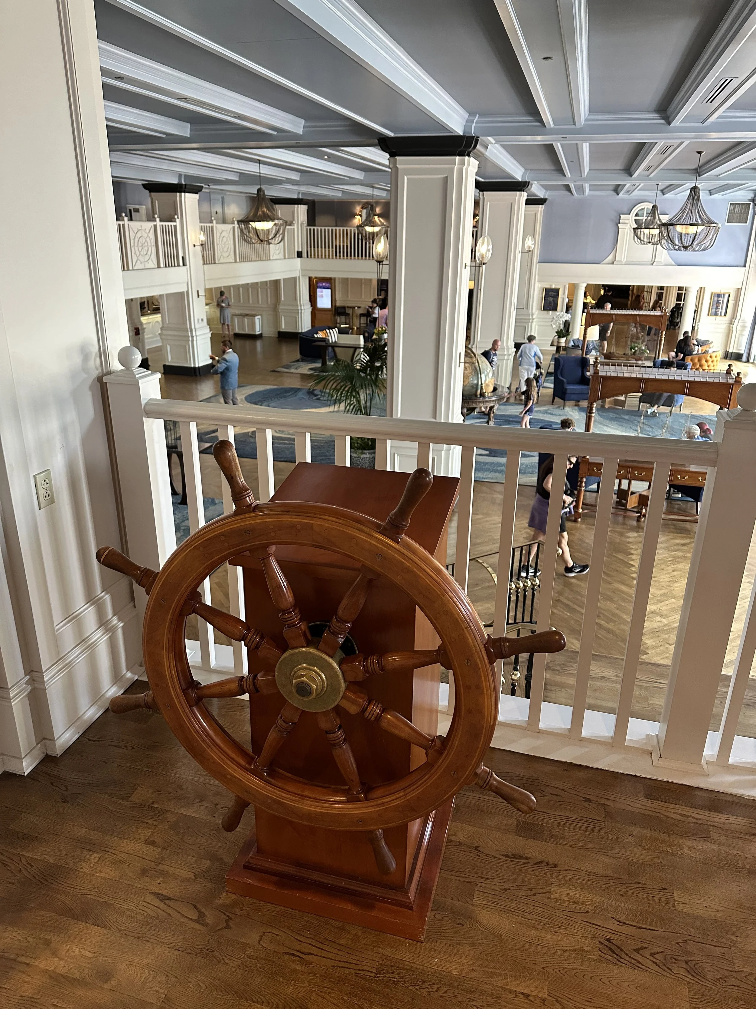 Interior of a hotel lobby with white columns, chandeliers, and people walking around. A wooden ship's wheel is positioned on a small stand near a white railing in the foreground.