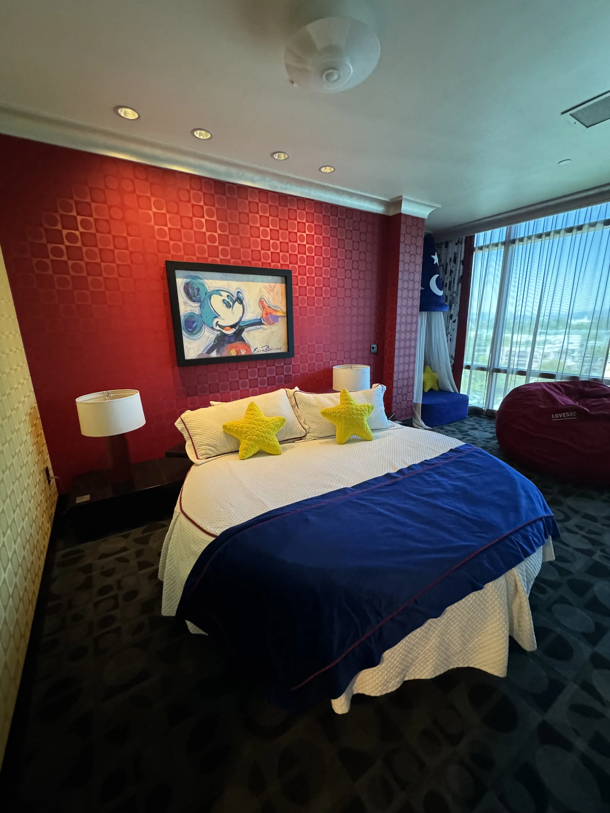 A hotel room with a red accent wall featuring a Mickey Mouse painting, a bed with white linens and three yellow star-shaped pillows, two white lamps on dark bedside tables, a large window with vertical blinds, and a purple bean bag chair on a pattern