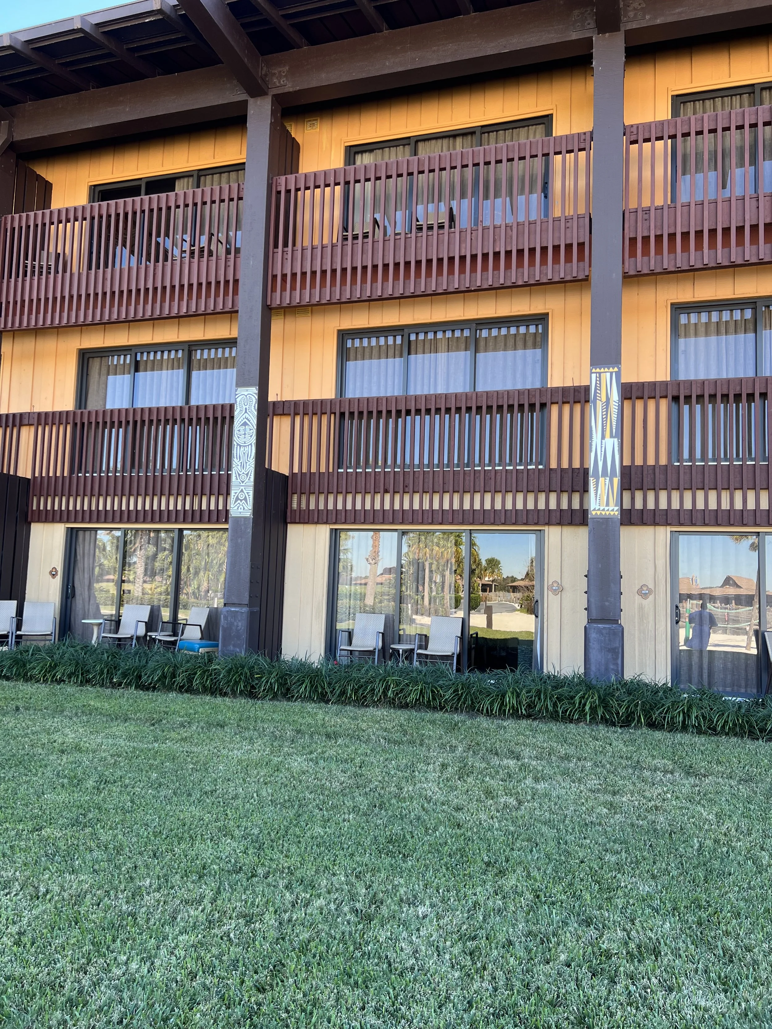 Exterior view of a multi-story building with wooden balconies, glass doors, and chairs on the ground level, surrounded by green grass and plants.