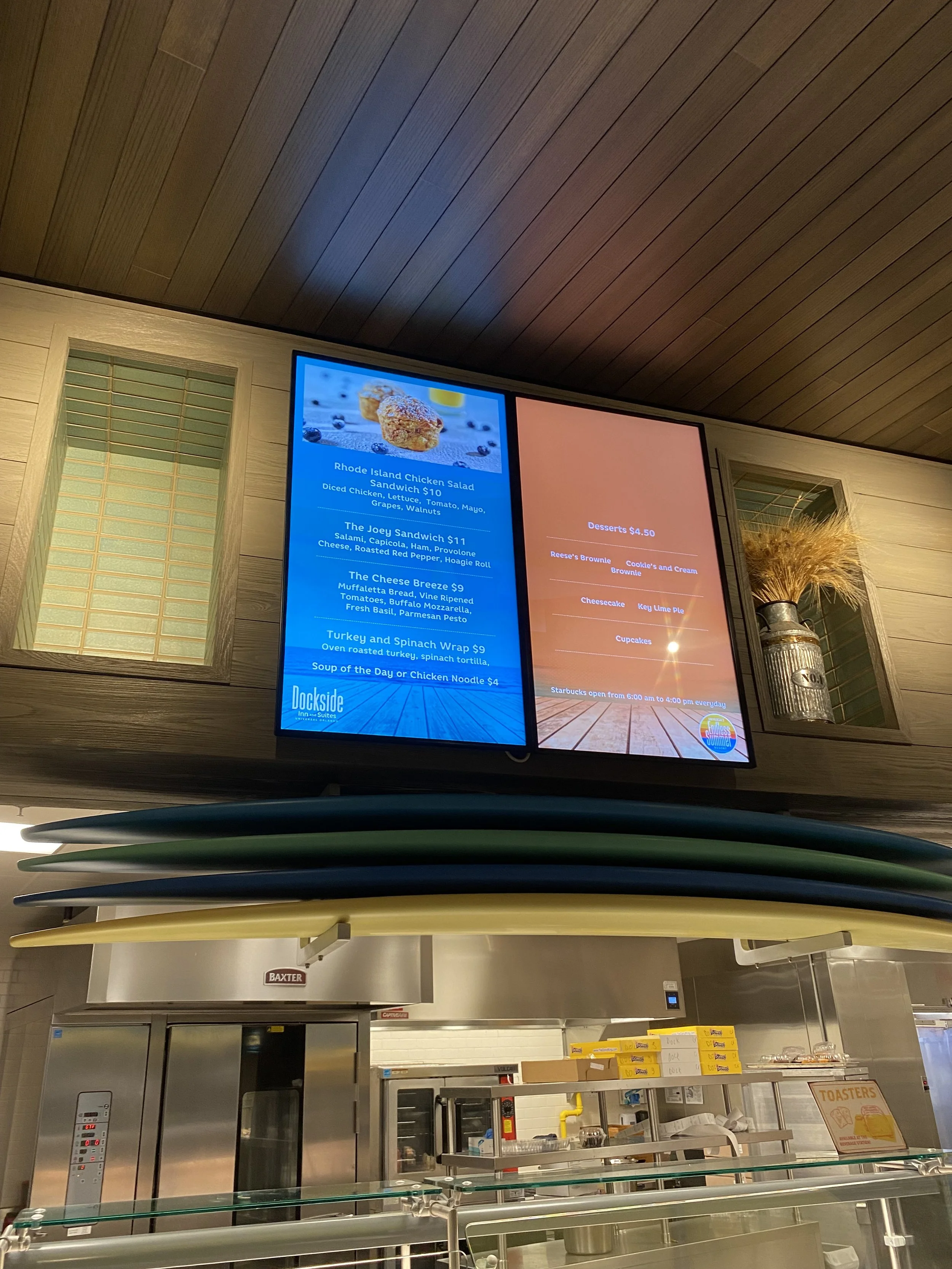 Digital menu screens displaying food options, with surfboards stacked underneath, in a restaurant or cafe setting.