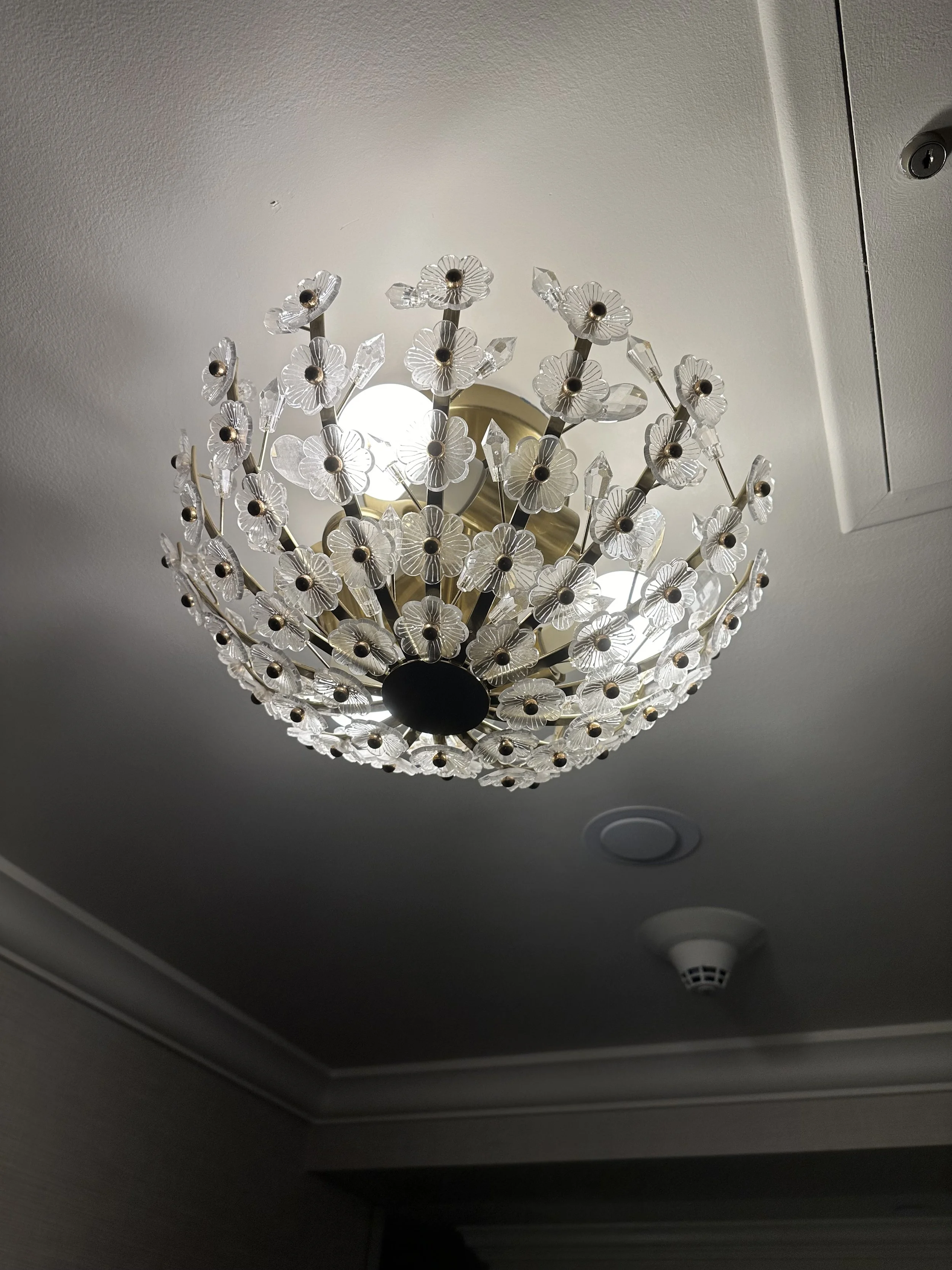 A decorative chandelier with glass flower-shaped elements and crystal accents hanging from a gold-colored metal base, mounted on a ceiling with white paint.