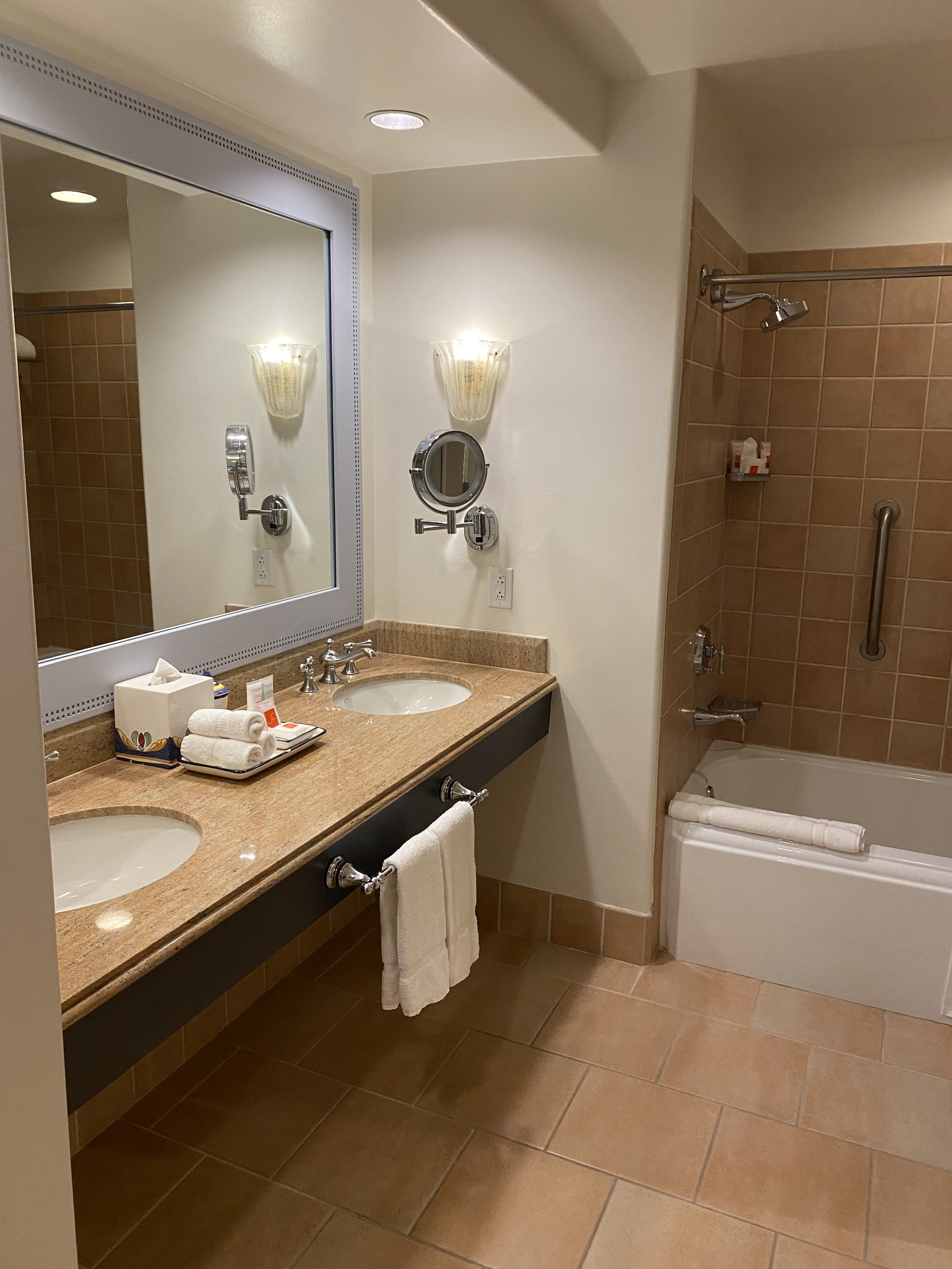 A hotel bathroom with a double sink vanity, large mirror, wall-mounted lights, a small round mirror, and a bathtub with a shower. The vanity has toiletries and towels, and the room features beige and brown tiles.