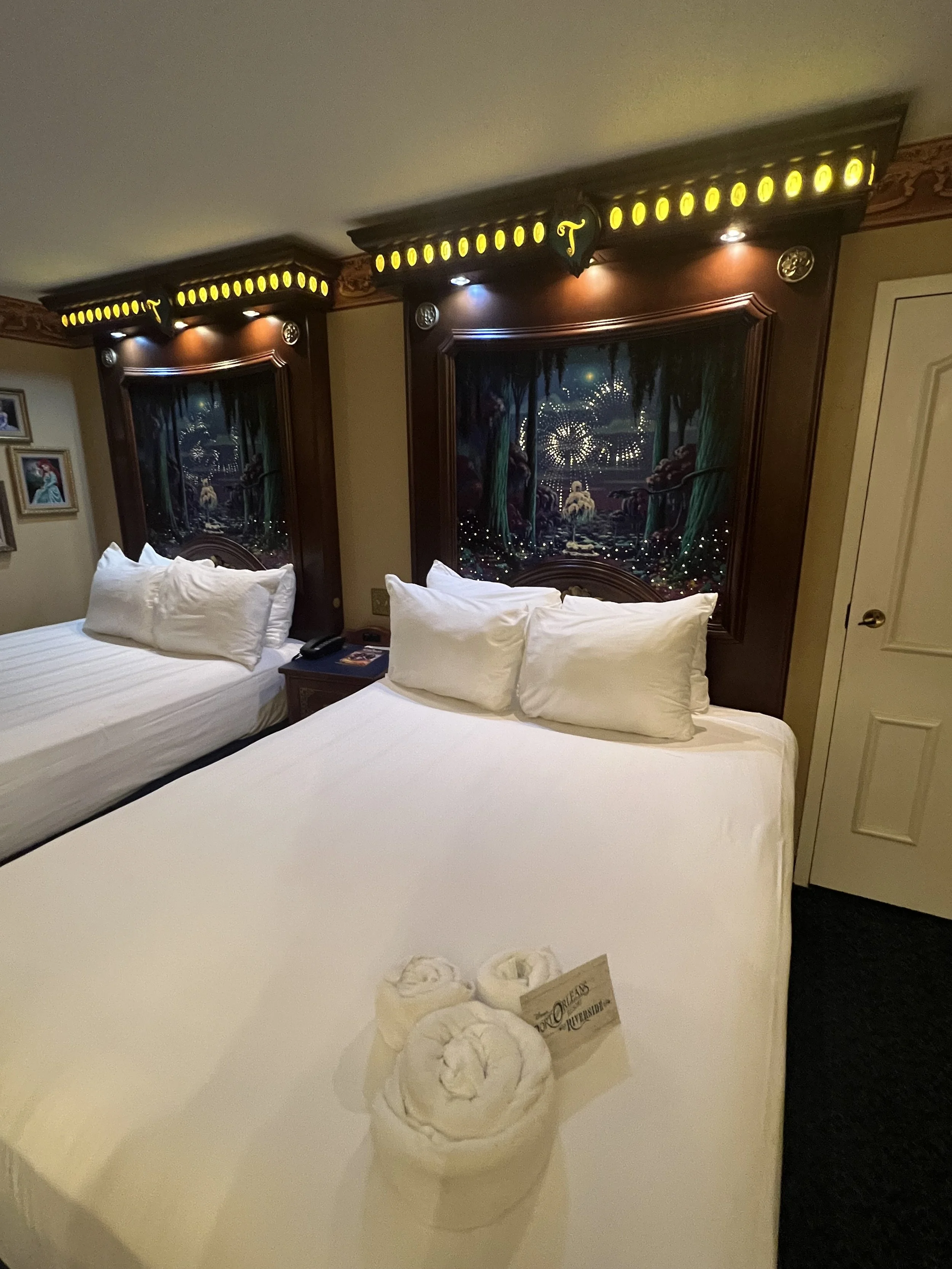 Two beds with white linens and pillows in a hotel room with themed headboards. Towels folded into animal shapes on the front bed, with a small paper sign. Artwork on the wall and a nightstand with a telephone and booklet.