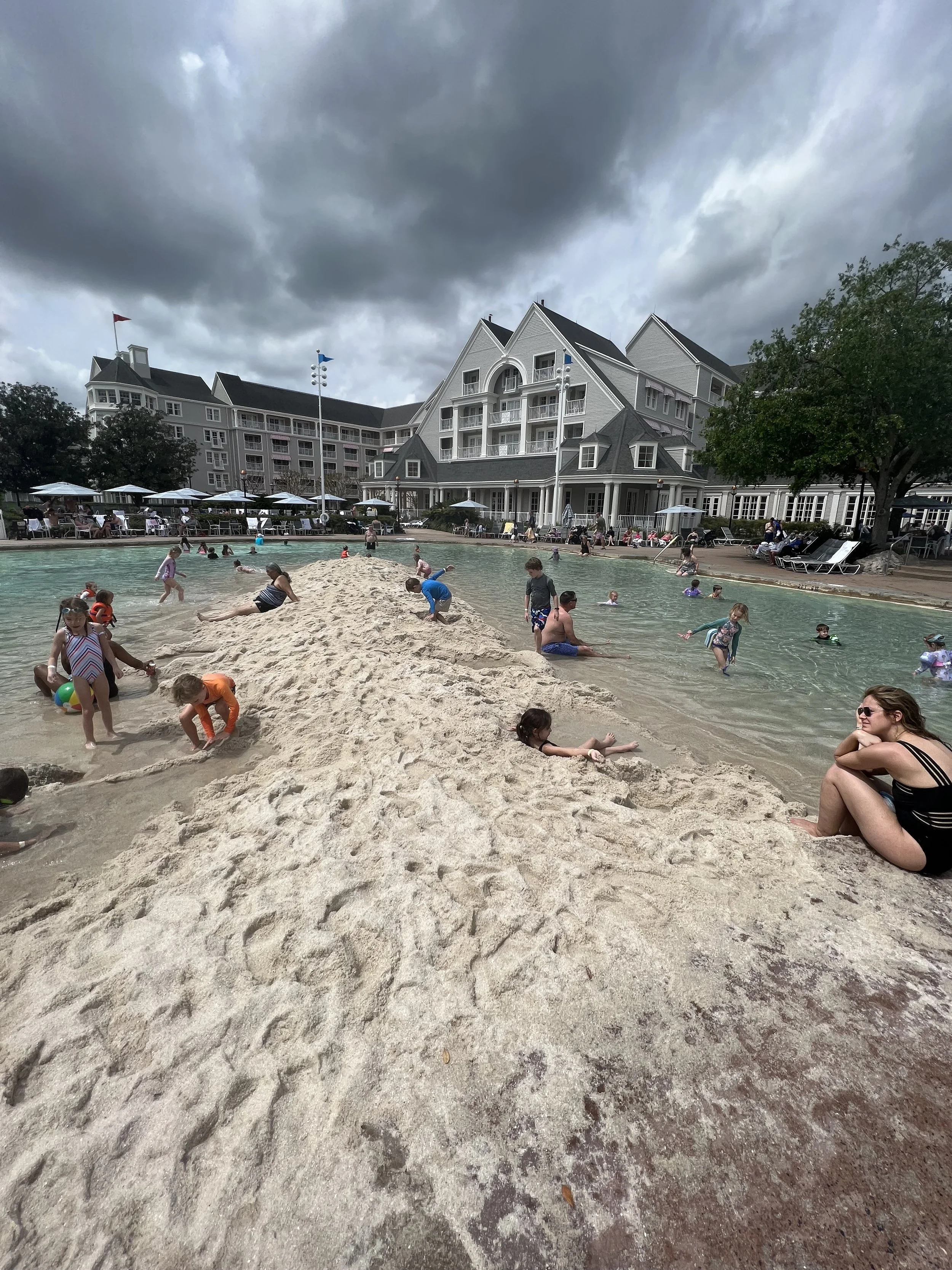 Children and adults playing and swimming at a beach with a sandbar, with a large hotel building and dark stormy clouds overhead.
