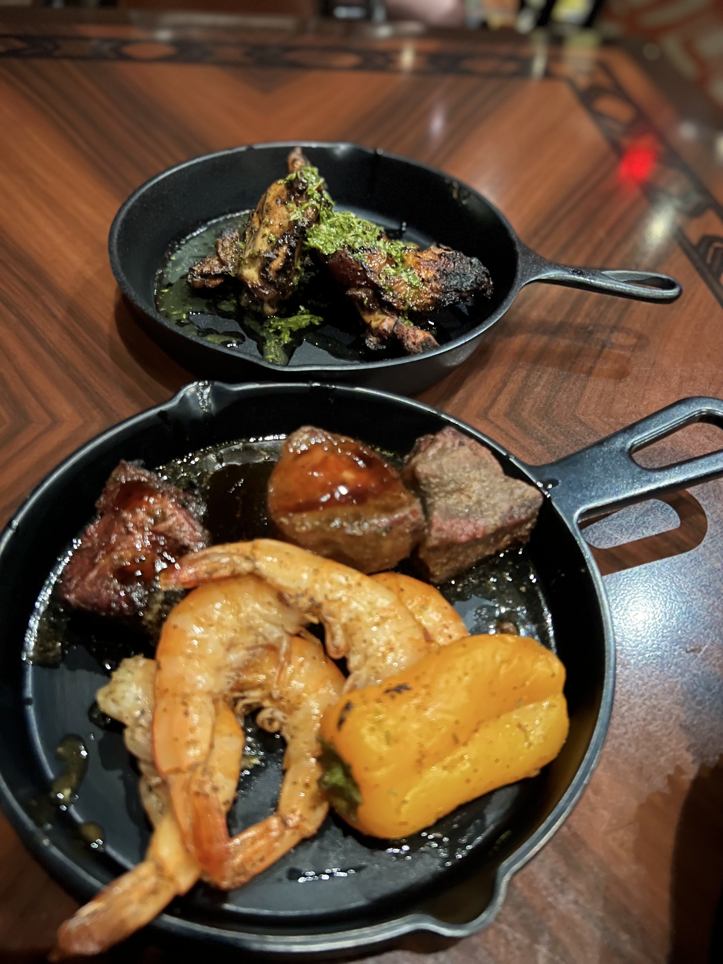 Cooked shrimp, steak, and yellow bell pepper on cast iron skillets, with cooked chicken wings and seasoned chicken pieces in the background.