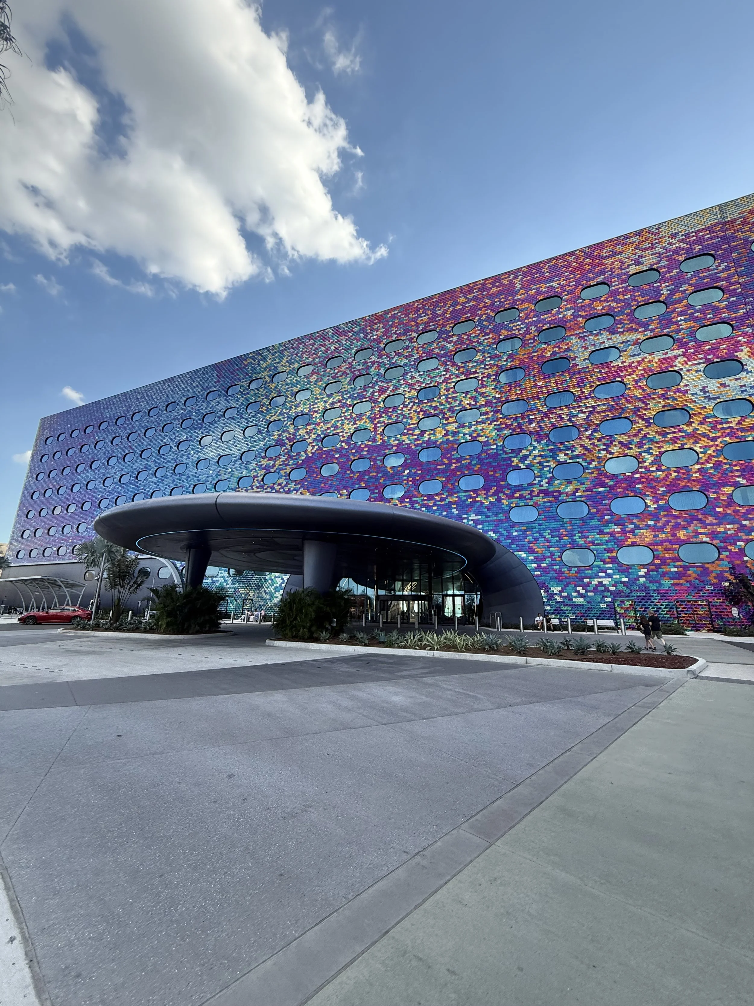 Colorful, modern building with many small, oval windows and a rounded entrance canopy, set against a blue sky with scattered clouds.